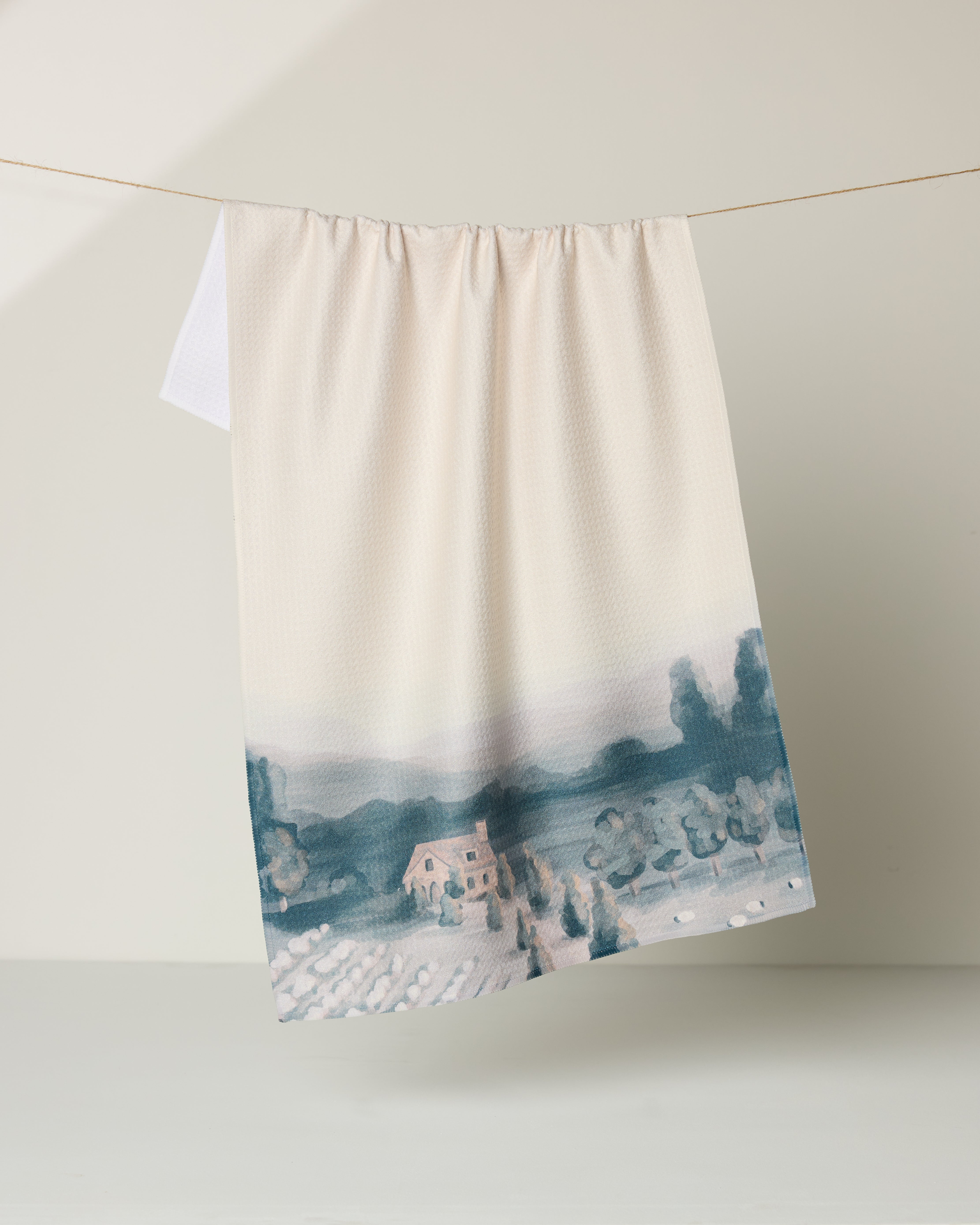The Hannah Langford "French Cottage" kitchen towel, cream-colored and eco-friendly with a blue and beige landscape design, hangs on a clothesline against a neutral background.