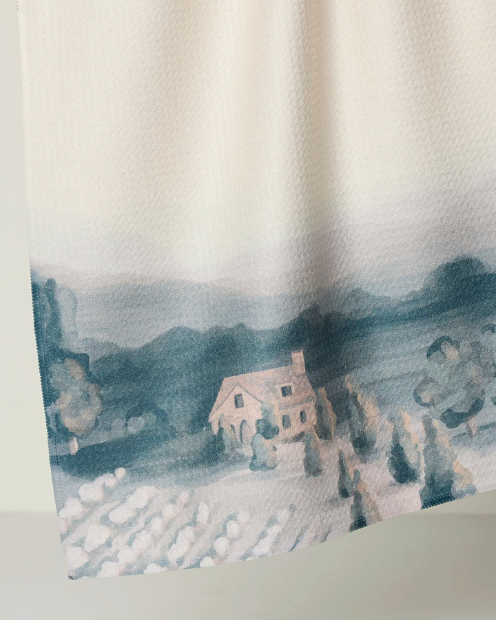A close-up of Hannah Langford’s French Cottage tea towel, cream-colored with a soft watercolor cottage scene among trees and fields, distant blue-gray hills, and thoughtfully crafted from recycled materials.