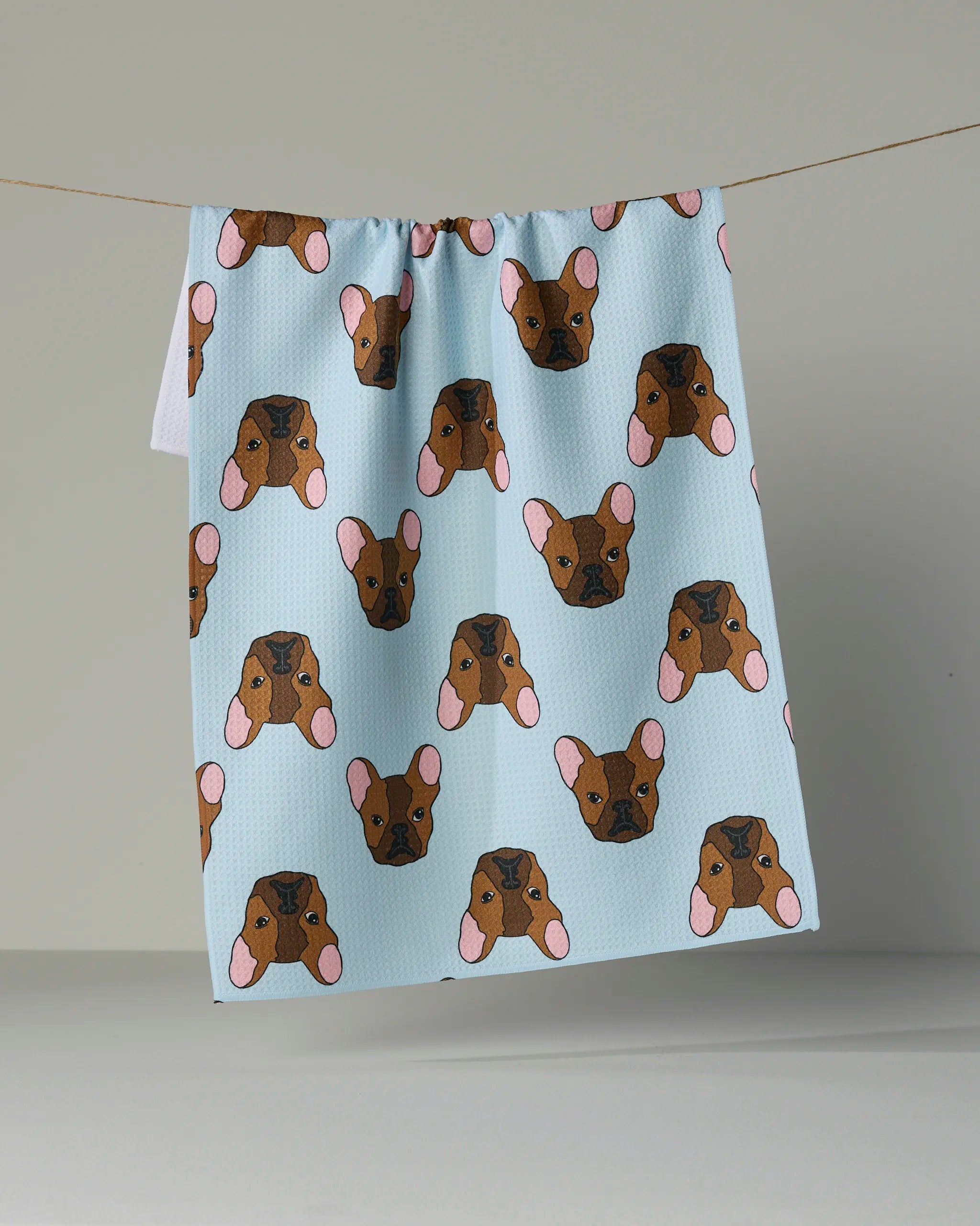 A light blue Geometry tea towel from the Frenchie Faces collection, made from recycled materials and featuring a repeating pattern of brown French Bulldog faces with pink ears, hangs on a line against a plain background.