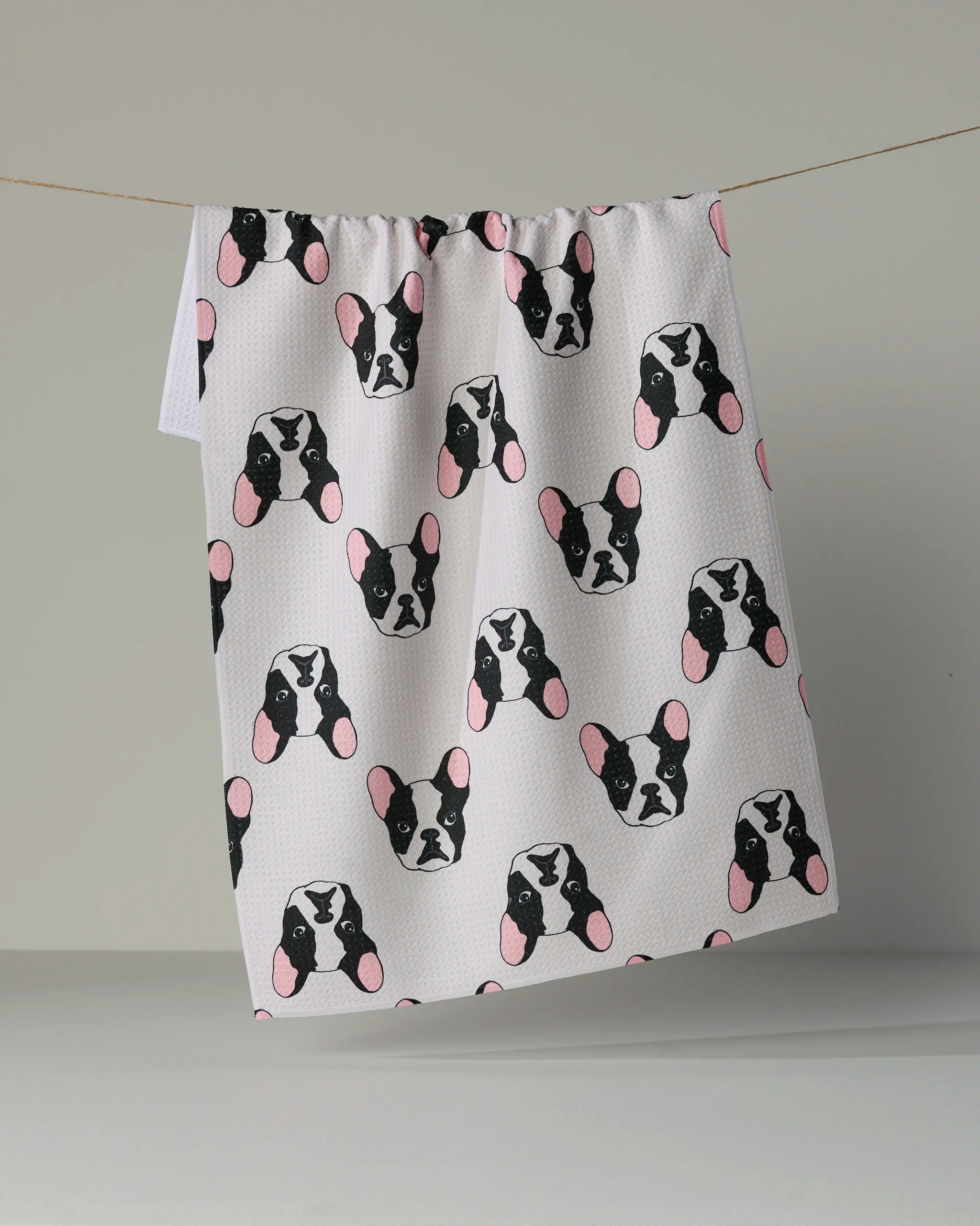 The Geometry “Frenchie Faces” kitchen tea towel features a playful, repeating pattern of black and white French bulldog faces with pink ears on a crisp white background. Made from recycled materials, this ultra-absorbent towel combines eco-friendly practicality with charming design.