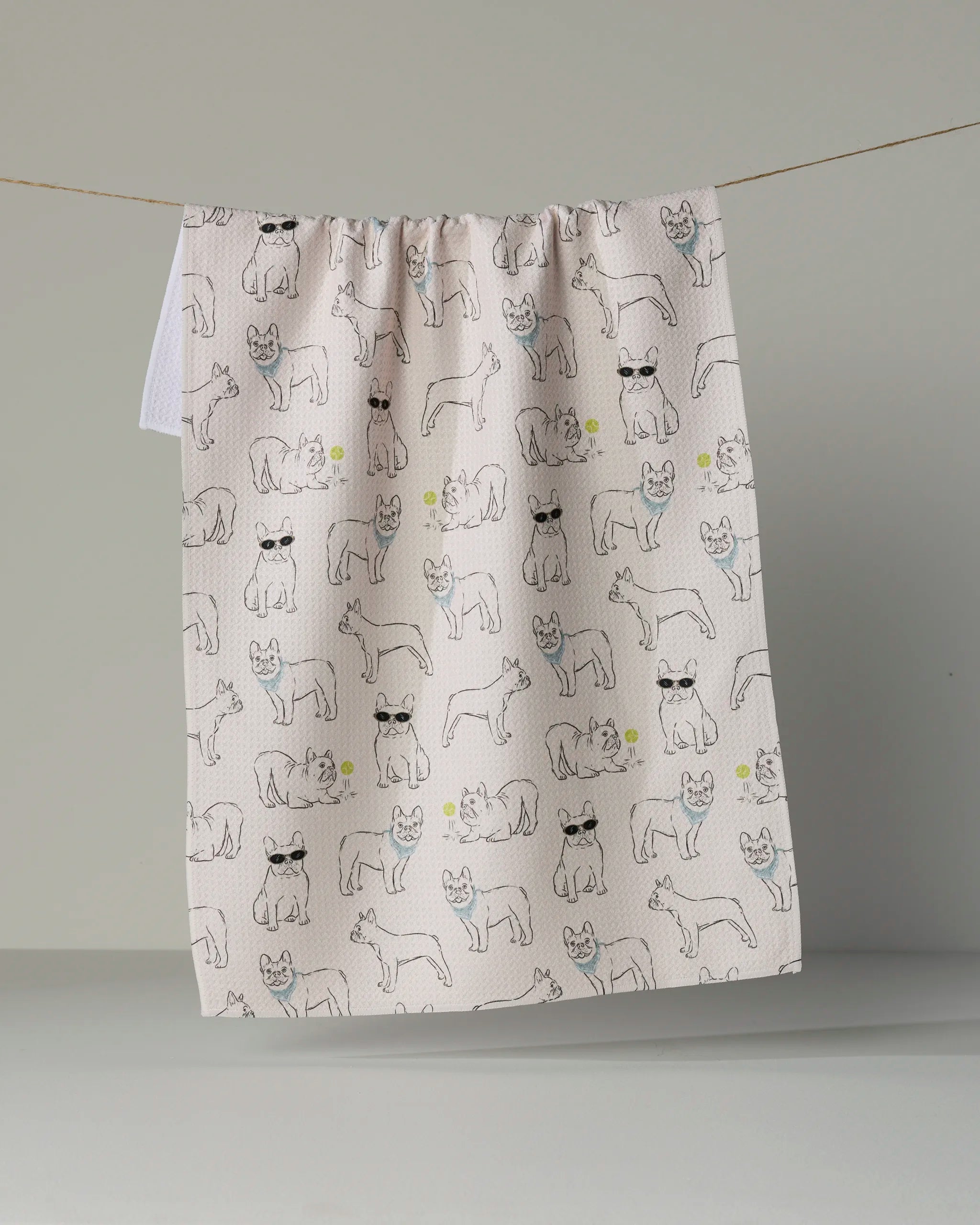 Brighten up your kitchen with the “Frenchie Frolic” tea towel from Geometry. Made from recycled materials, this light-colored towel features a playful pattern of outlined Frenchies—some sporting sunglasses—intertwined with small yellow flowers. Hanging against a plain gray background, it adds both sustainability and whimsical charm to your home.