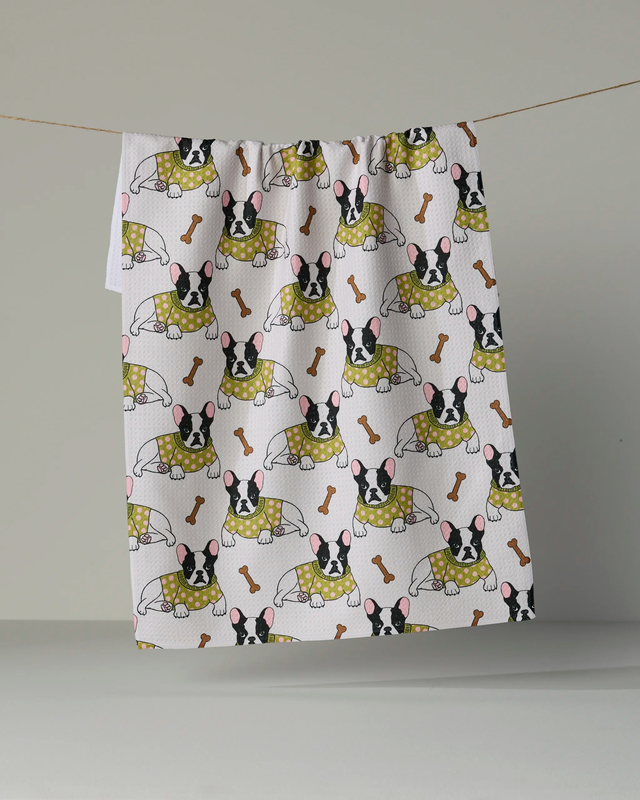 A tea towel from Geometry’s Frenchie Fun collection hangs on a line, showcasing a playful pattern of French bulldogs in yellow outfits with brown bones scattered across a crisp white background.