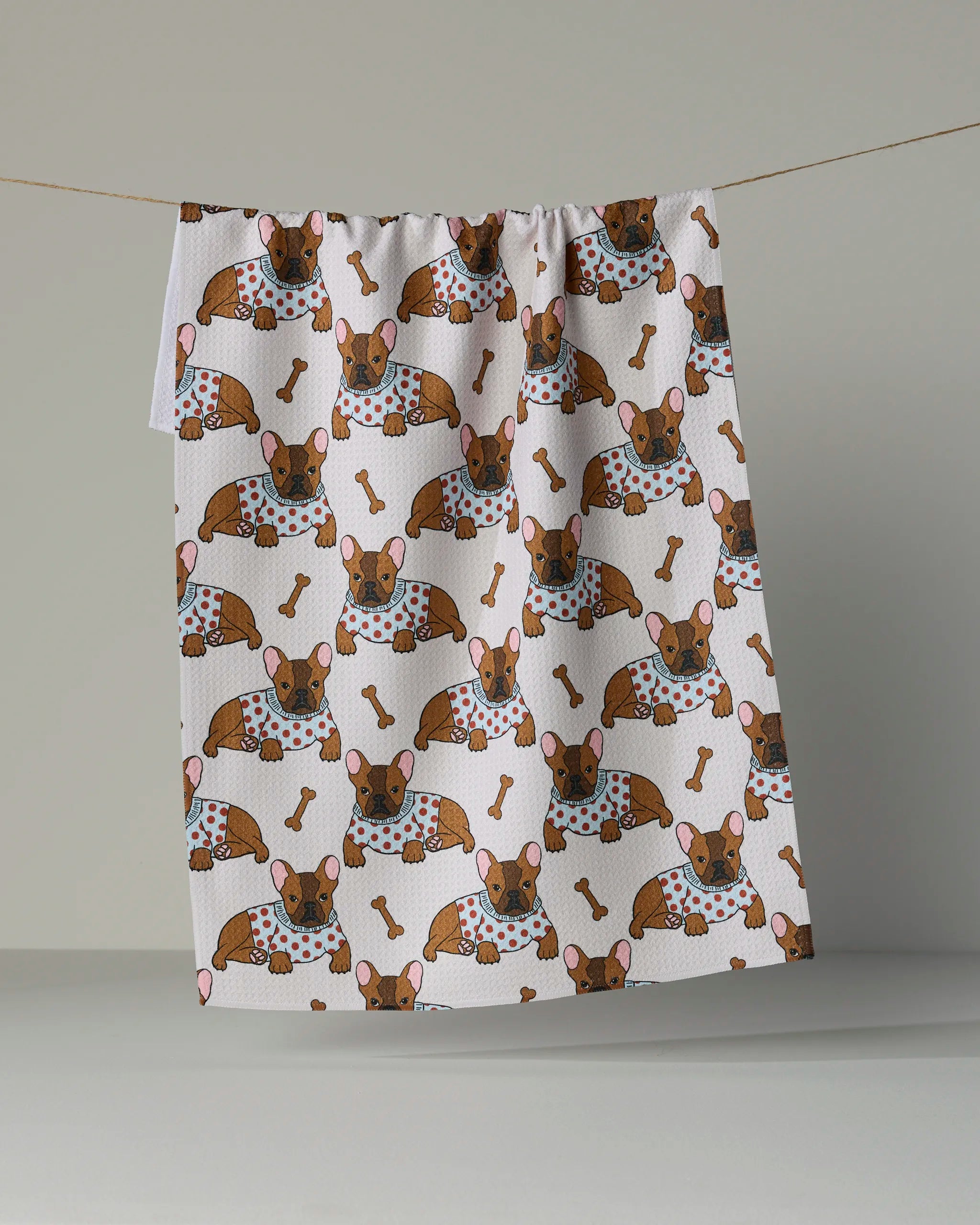 A white tea towel from Geometry, called “Frenchie Fun,” hangs on a line, showcasing a repeating pattern of cartoon brown dogs wearing polka-dot bibs and surrounded by bones.