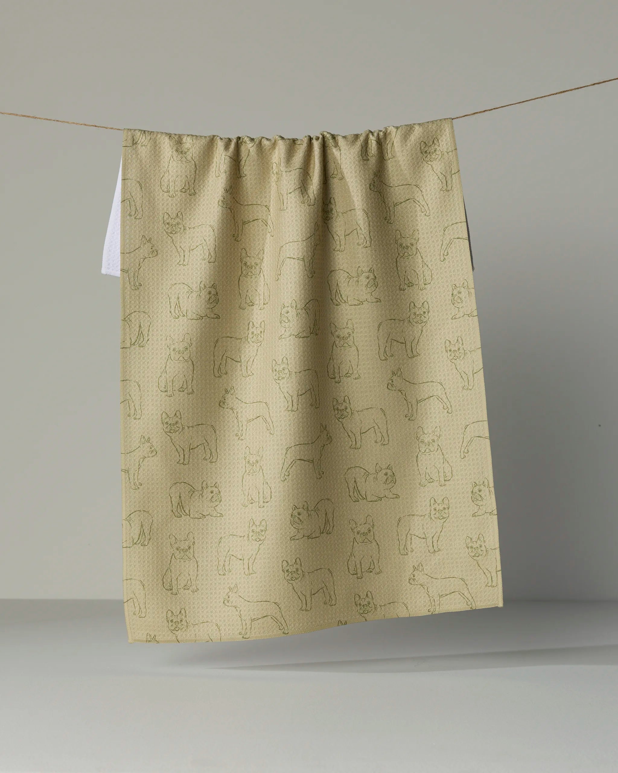 A tea towel from Geometry called “Frenchie Pups,” featuring a simple outlined French bulldog pattern in beige, hangs on a string against a plain background.