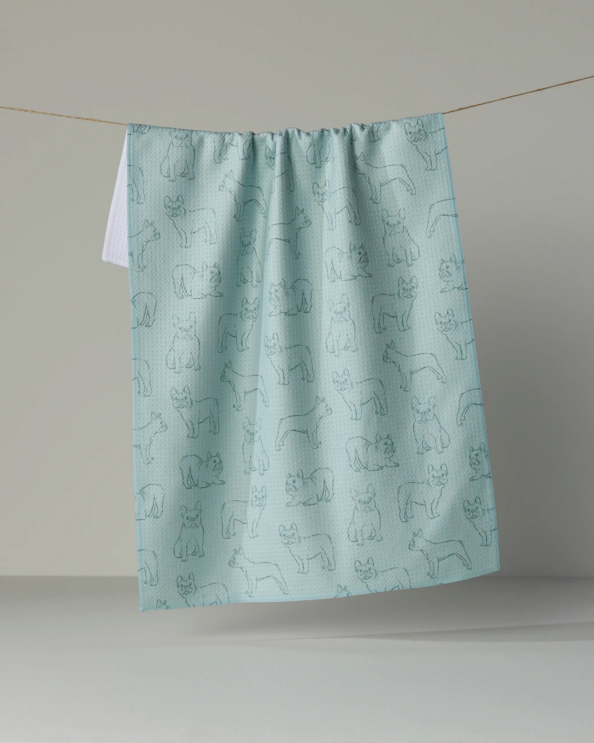 A light blue absorbent dish towel from Geometry, featuring an outline pattern of various Frenchie pups, hangs on a clothesline against a plain light-colored background.