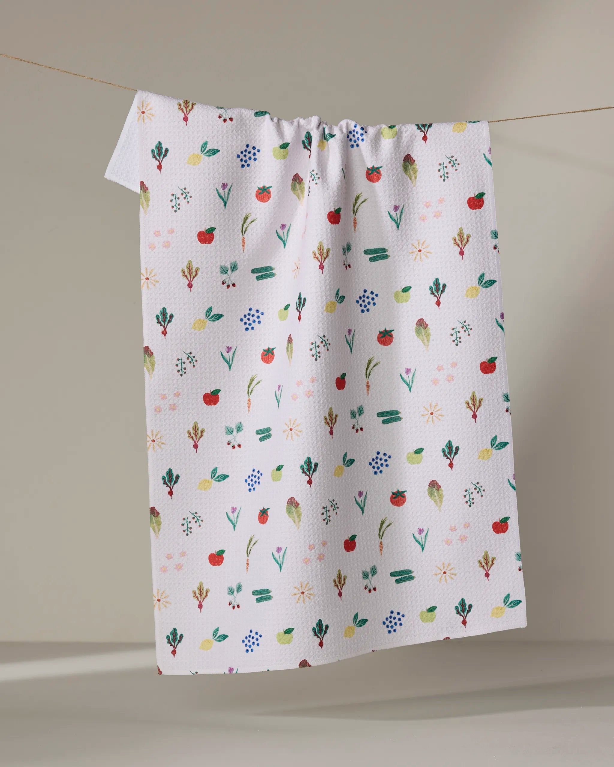 The Fresh Produce dish towel by Brittany Jepsen, featuring a colorful pattern of fruits, flowers, and leaves on white fabric, hangs on a clothesline against a neutral background.