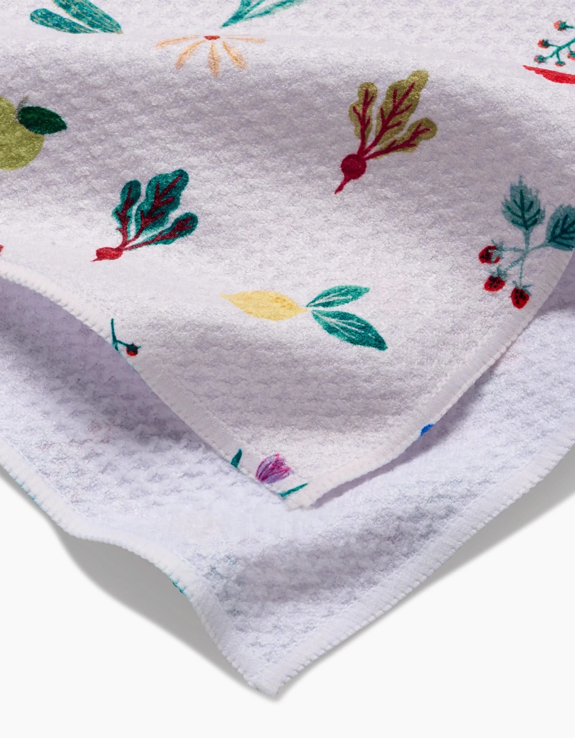 The Fresh Produce tea towel by Brittany Jepsen features colorful embroidered veggies and leaves on a white textured Geoweave fabric, offering extra absorbency and softness. Shown folded at the corner.