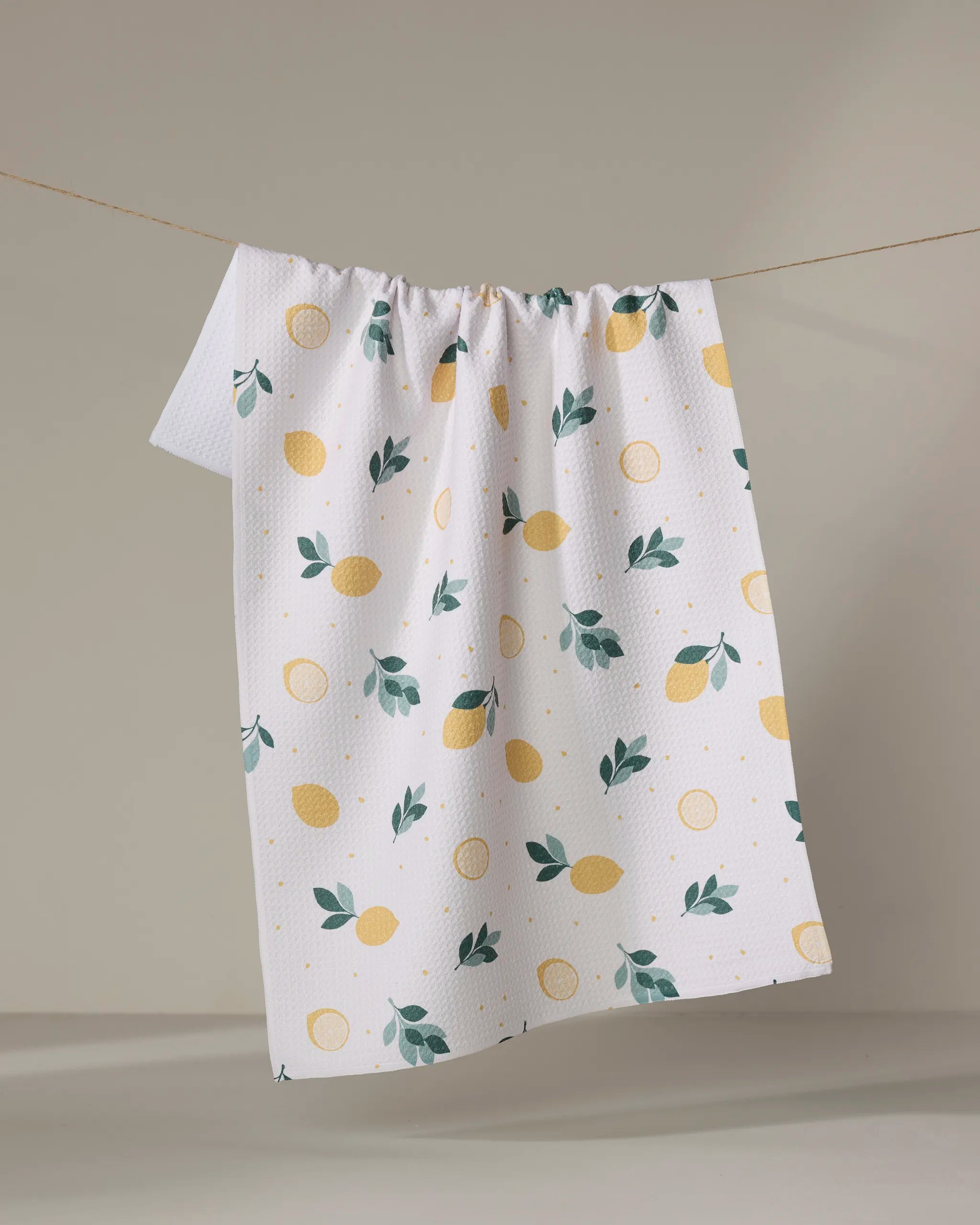 The "Freshly Picked" tea towel by Sara Mari, featuring a yellow lemon and green leaf pattern on white textured recycled fabric, hangs on a string against a light background.