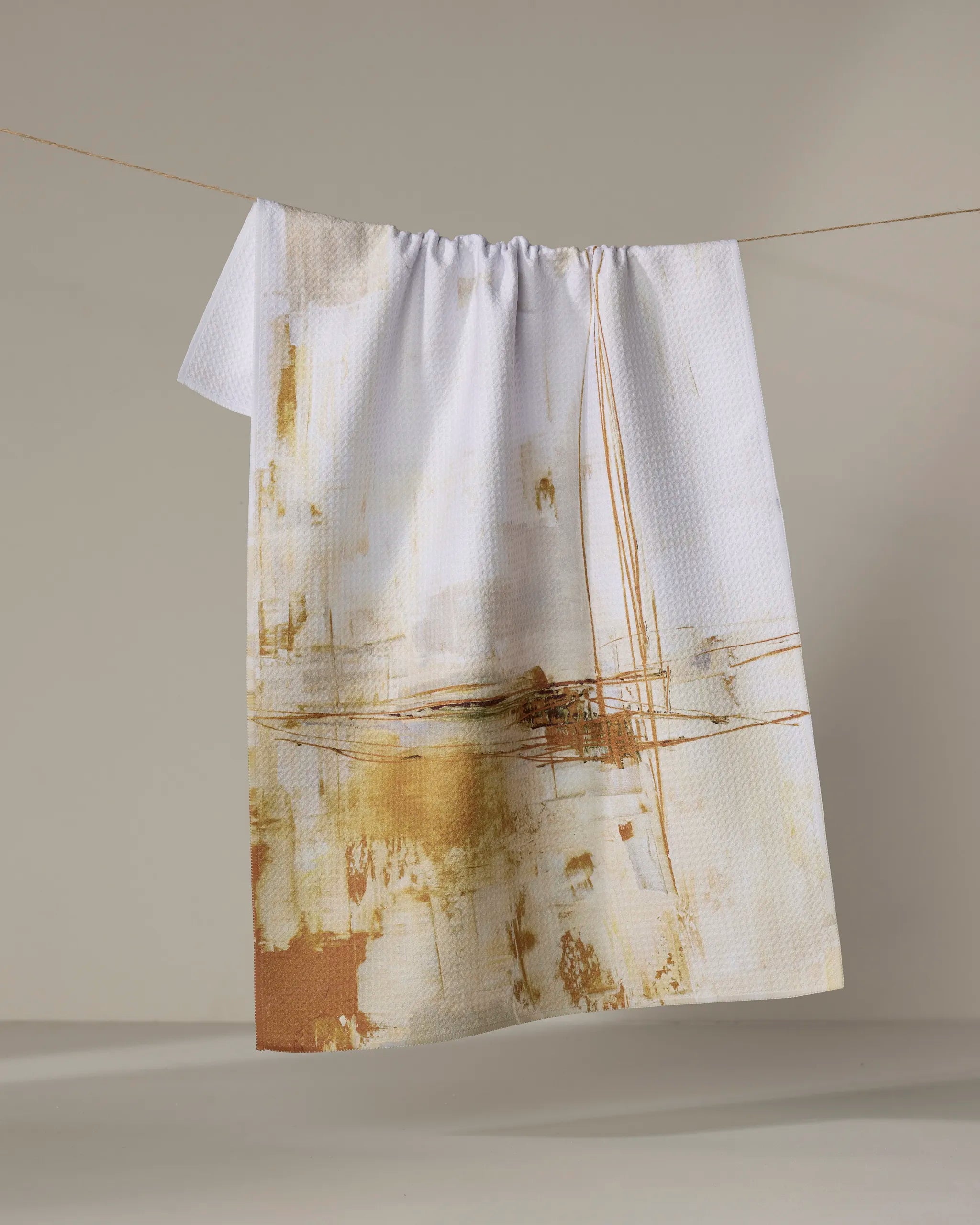 The Friction tea towel by Jennifer Janesko, crafted from recycled materials, features an absorbent design with an abstract gold and beige pattern and hangs from a clothesline against a neutral background.
