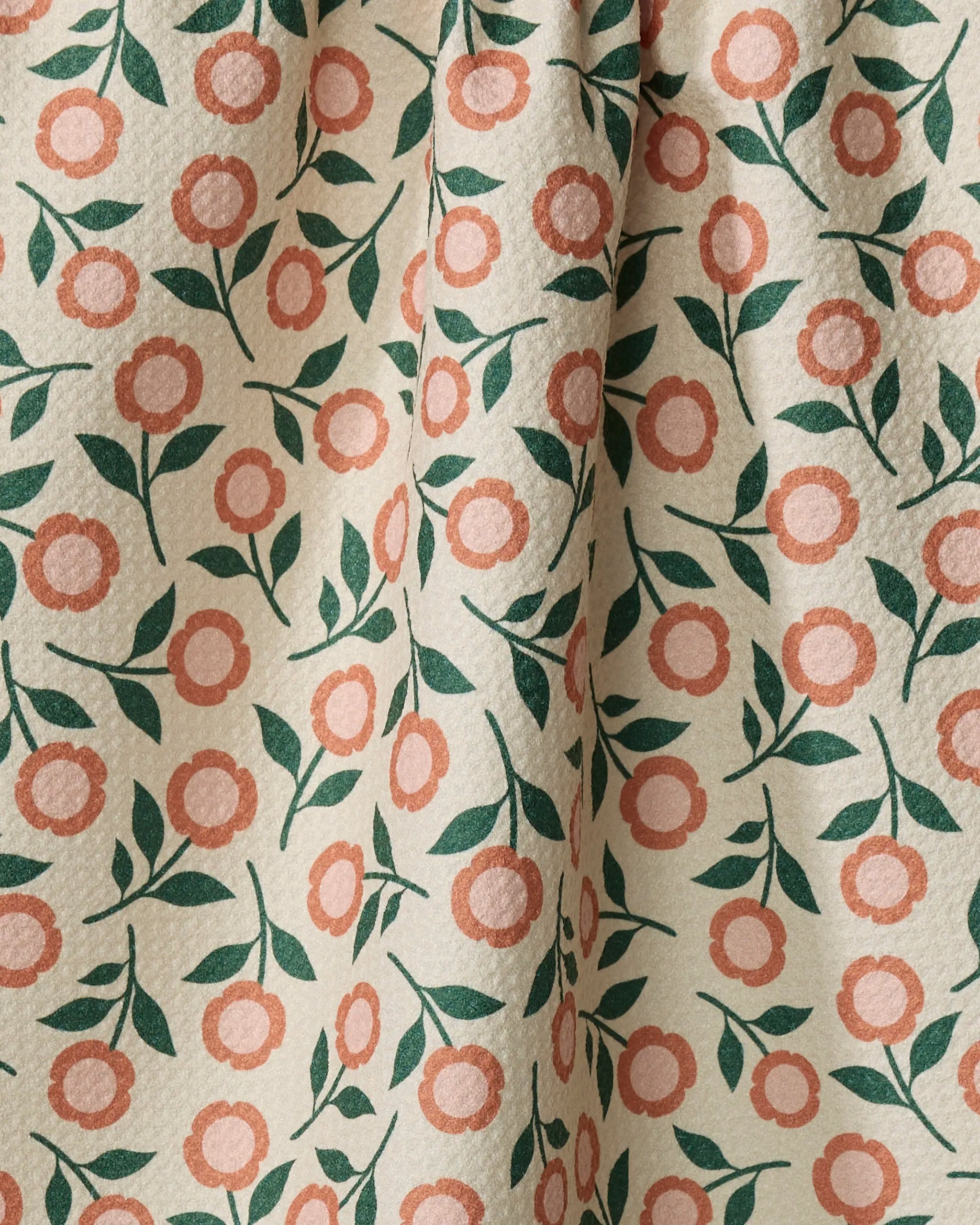 A close-up of Megan Kampa's Friendly Floral Peach tea towel, made from recycled materials, shows pinkish-orange flowers and green leaves on cream fabric with a textured surface and gentle folds. Ultra-absorbent and eco-friendly.
