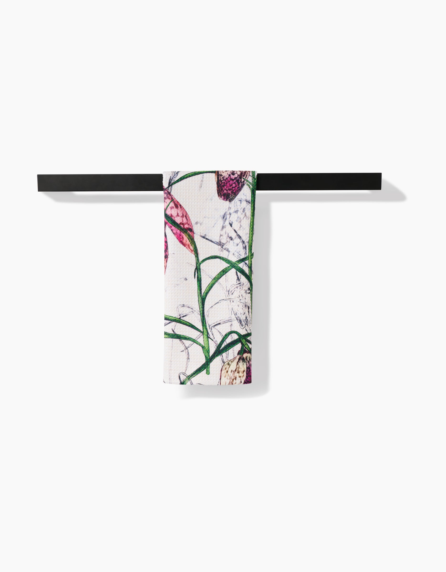 The Fritillaria tea towel by Pascale Dilger, made from recycled materials, showcases green leaves and purple flowers on white fabric and hangs neatly on a black towel bar against a plain white background.
