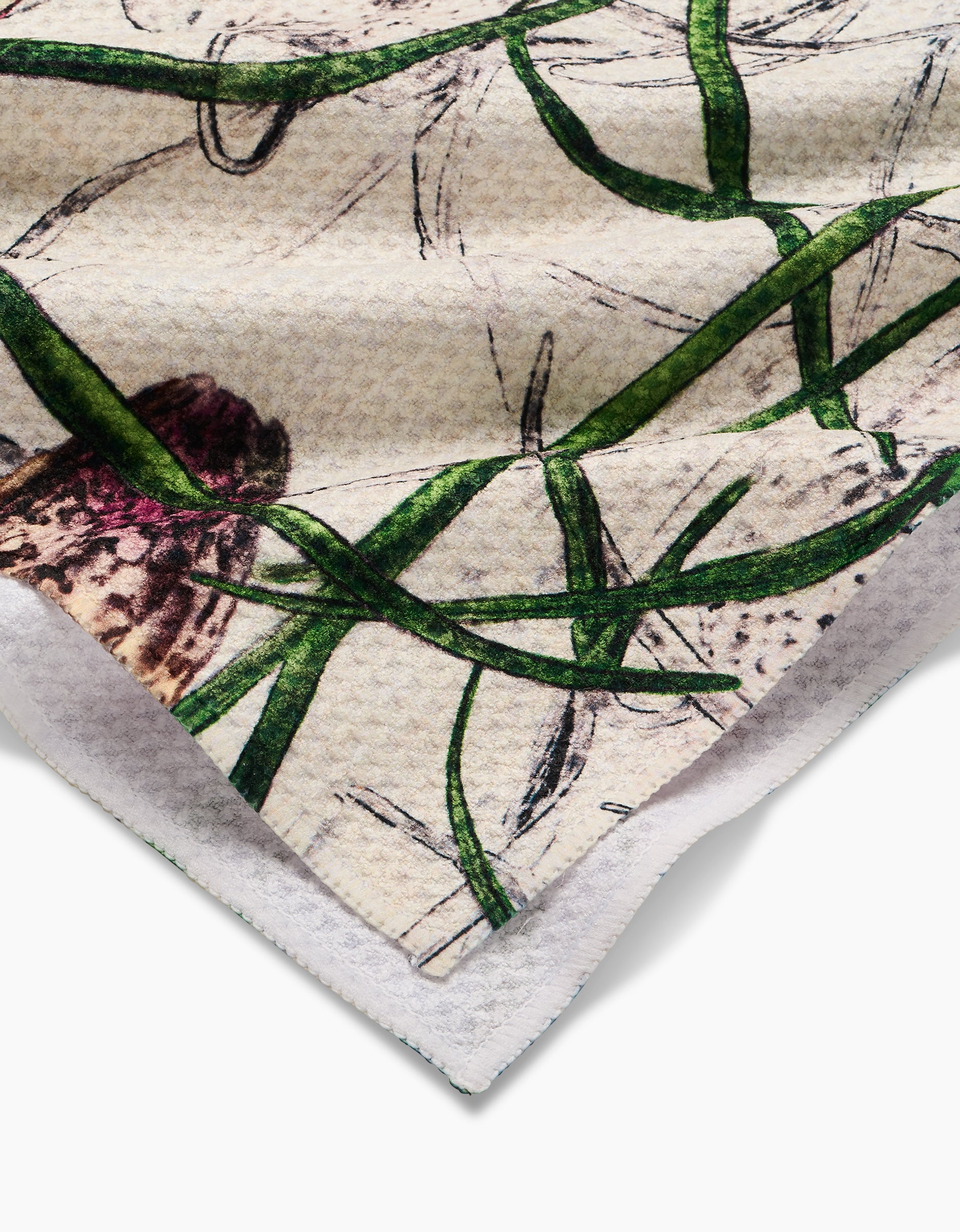 A close-up of Pascale Dilgers Fritillaria tea towel shows soft Geoweave fabric with green stems, leaves, and a touch of purple on cream. The towel is slightly folded, revealing its white underside.