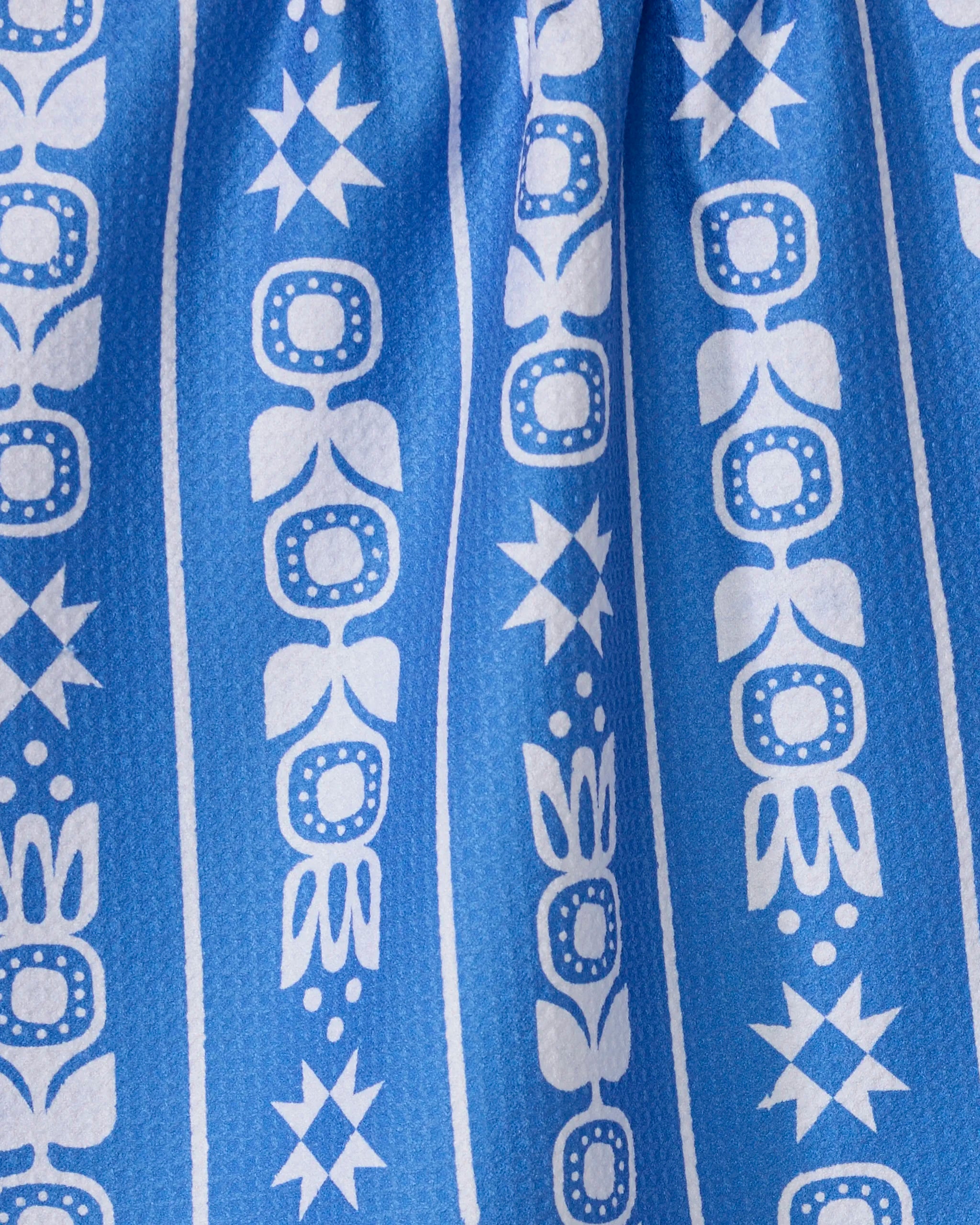 The Frosted Flora kitchen towel by Geometry features blue recycled fabric with vertical white geometric and floral patterns—stars, dots, and stylized leaves—in evenly spaced stripes. The top of the towel is gathered.