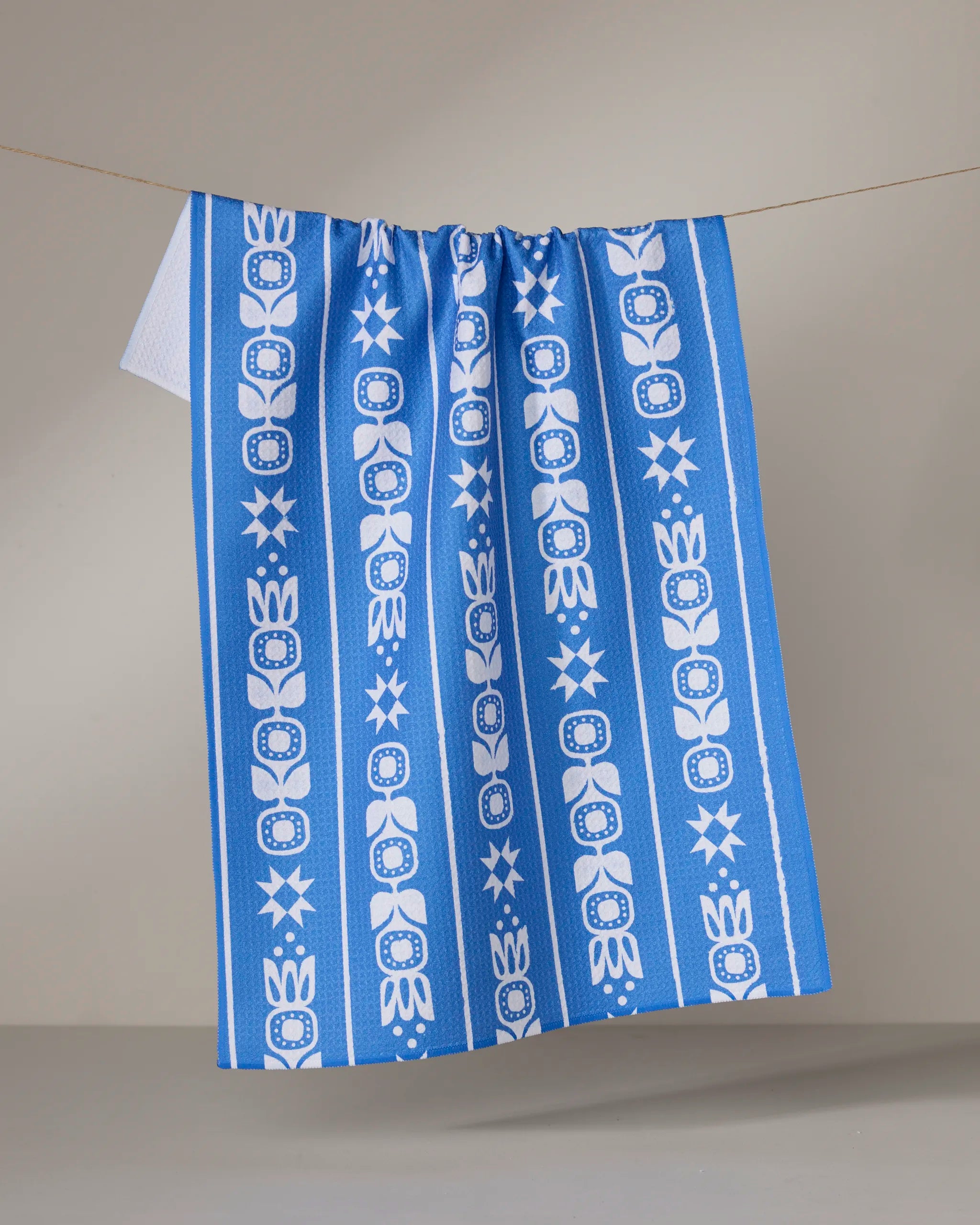 The Geometry "Frosted Flora" tea towel, in blue and white, hangs from a string. It features vertical stripes with floral and star motifs against a plain, neutral background.