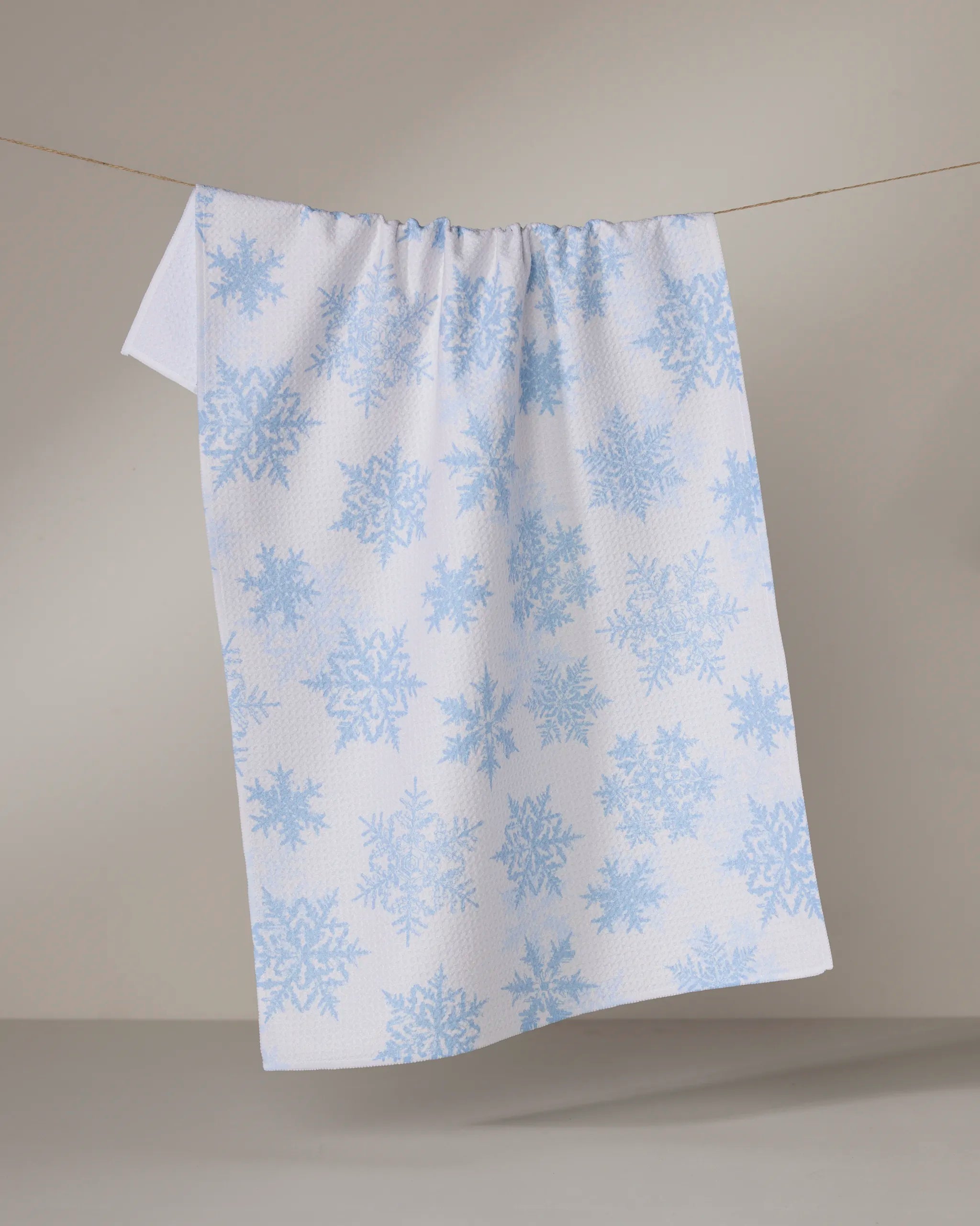 The Geometry Frostin kitchen towel, featuring light blue snowflake patterns on white and made from recycled materials, hangs on a clothesline against a neutral background.