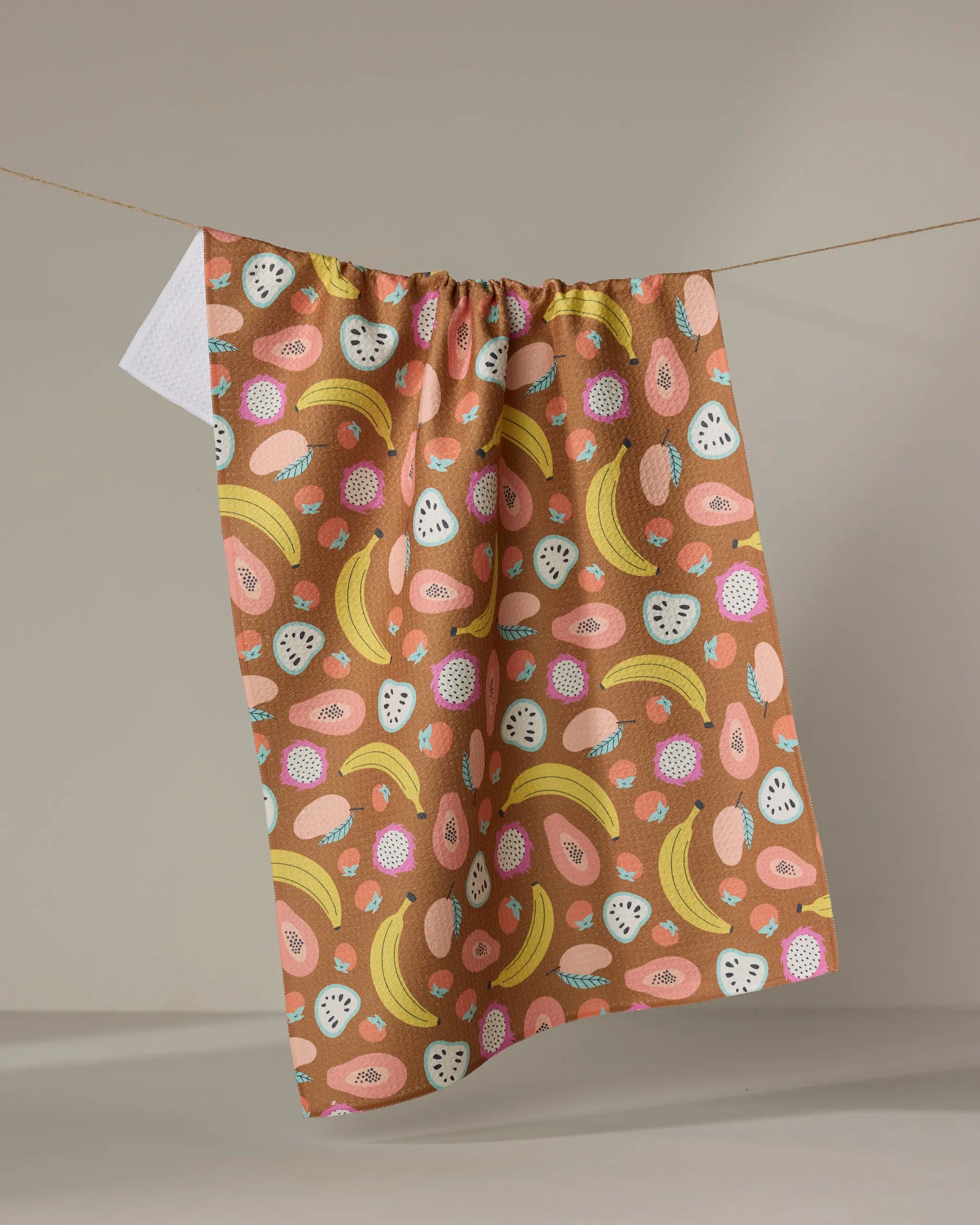 The Megan Kampa "Fruit Salad" kitchen towel features a vibrant pink, yellow, and white fruity pattern with bananas and dragon fruit, made from recycled materials and displayed on a line against a neutral background.