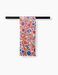 The Claire Iglesias Fruits Party tea towel, featuring a colorful abstract fruit and floral pattern in red, blue, white, and purple, hangs neatly from a black bar against a white background.
