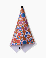 The Fruits Party tea towel by Claire Iglesias is brown, made from recycled materials, and features a colorful fruit pattern with red, blue, and green shapes. It hangs from a black loop against a white background.