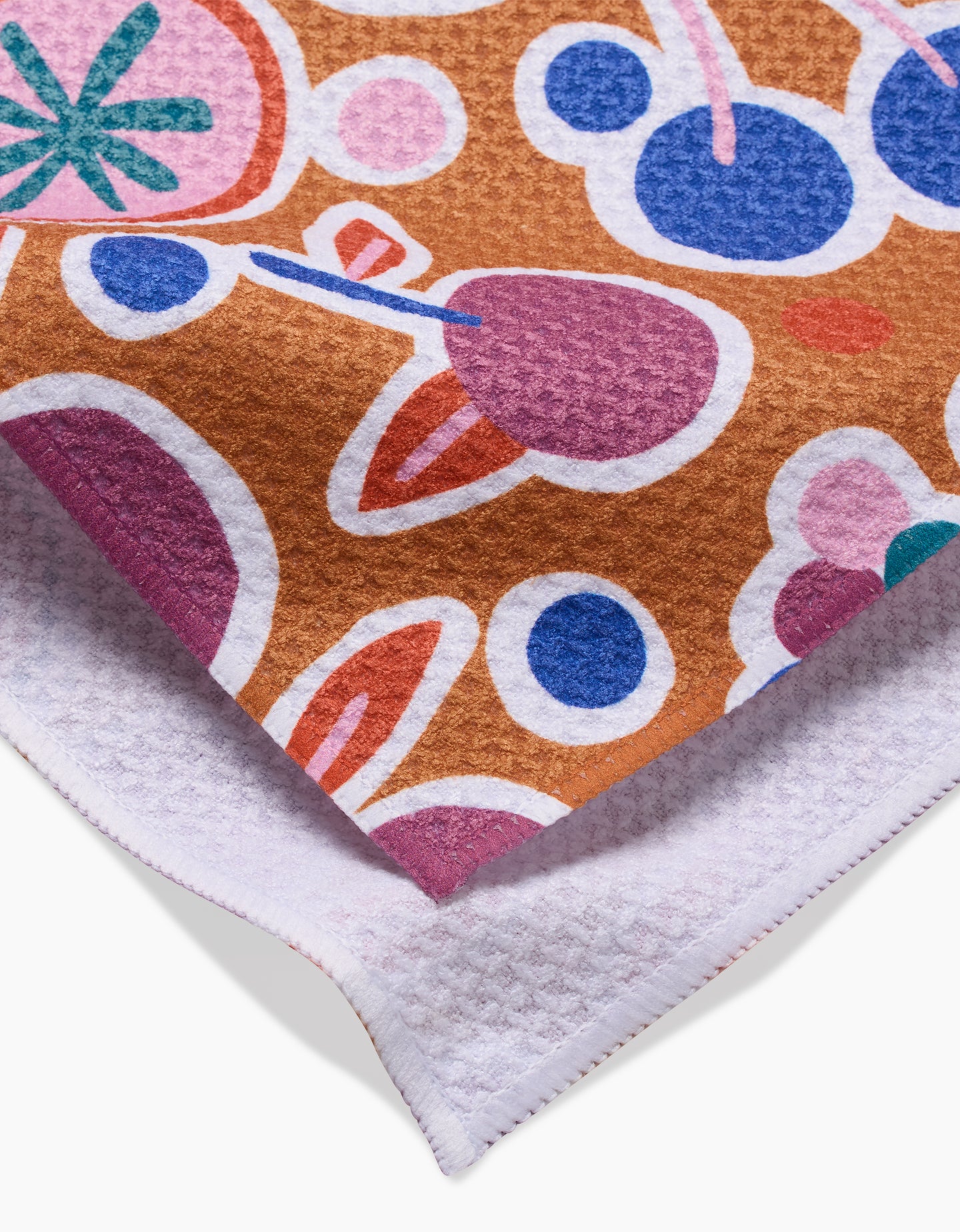 A close-up of Claire Iglesiass Fruits Party tea towel—ultra-absorbent, with a colorful abstract fruit pattern in pink, purple, blue, and green on brown. Features a textured white underside and two overlapping corners.