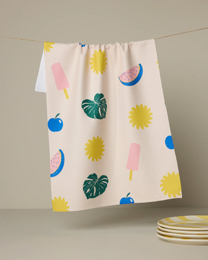 The Geometry Fruity Pop tea towel, featuring yellow flowers, green leaves, pink popsicles, blue apples, and pink watermelon slices, hangs on a line. Stacked plates with yellow stripes sit nearby beside this ultra-absorbent towel.
