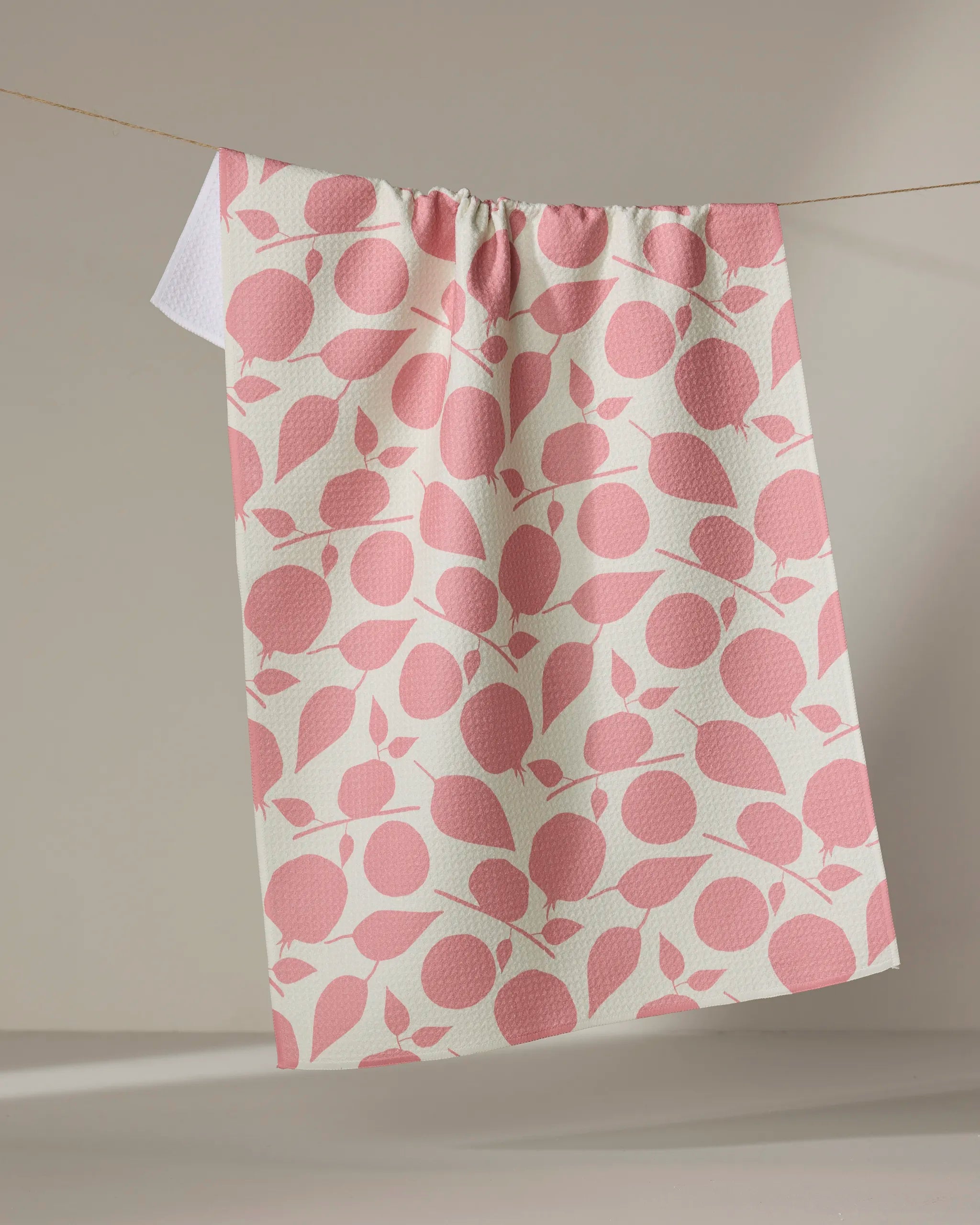 The Lizzie Clark Fruity Toss tea towel, featuring a pink fruit and leaf pattern, hangs on a string against a neutral background. Its quick-drying fabric makes it both stylish and practical for any kitchen.
