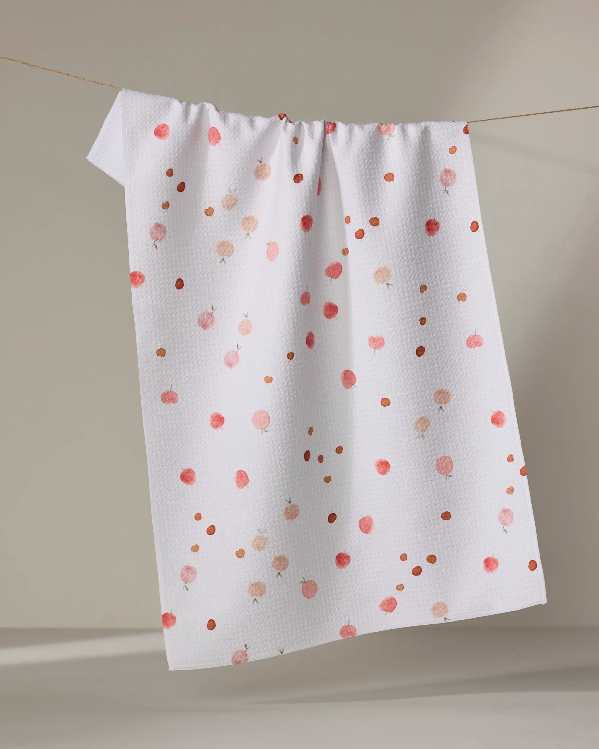 The Fruity tea towel by Anee Shah features a white textured design with pink and orange dot patterns, made from recycled materials, shown hanging on a clothesline against a neutral background.