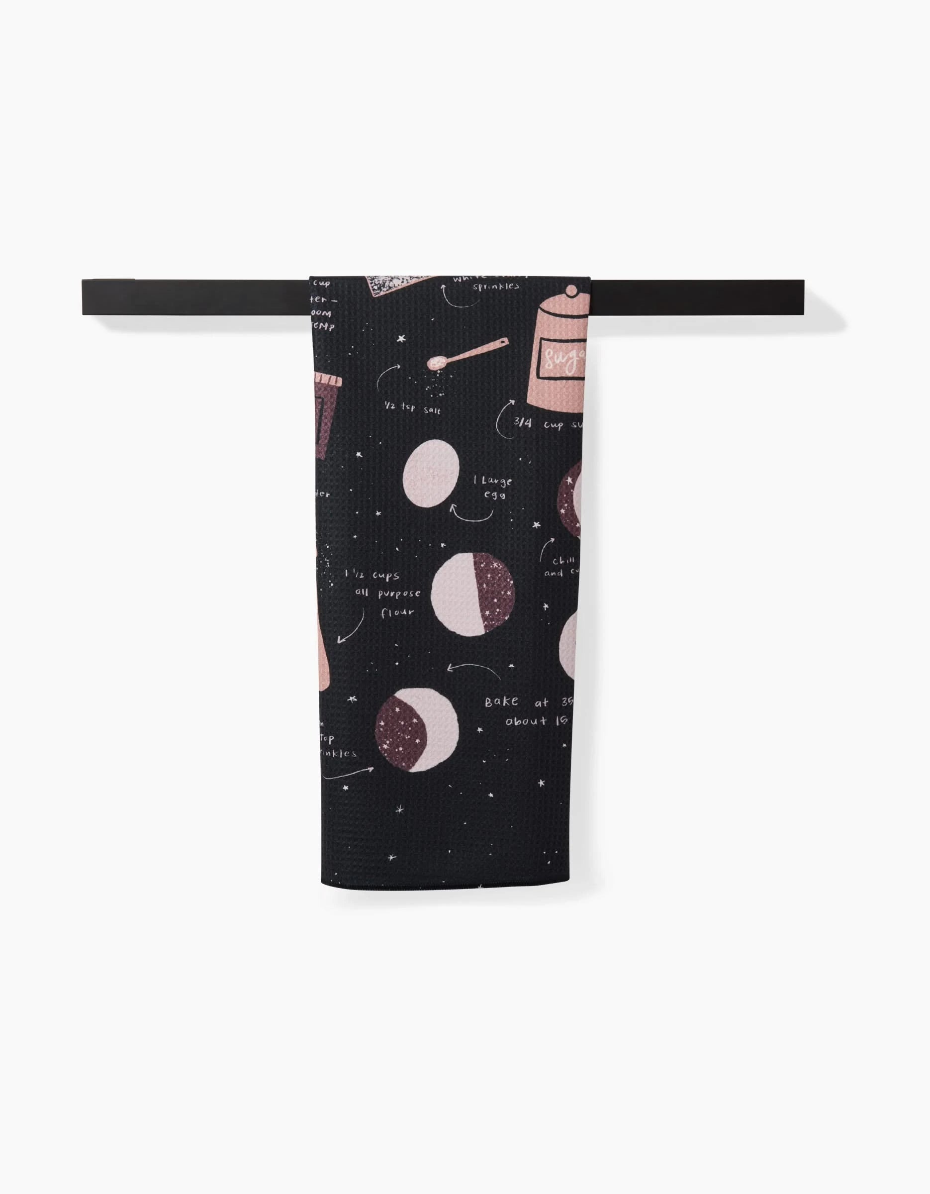 The Full Moon Cookies tea towel by Janna Sue Design, featuring eco-friendly black fabric with white and pink baking illustrations and instructions, is shown neatly folded on a horizontal bar against a plain white background.