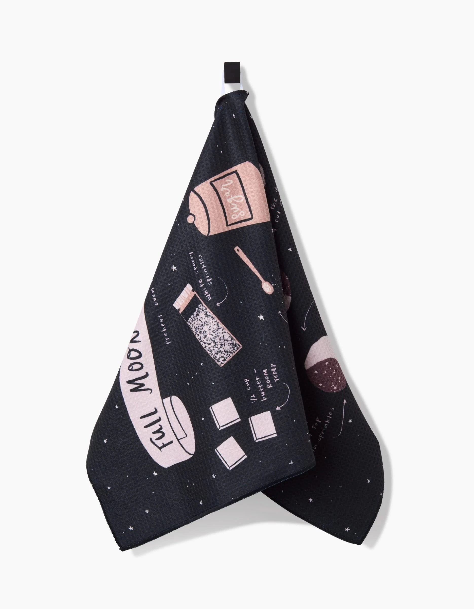 The Full Moon Cookies tea towel by Janna Sue Design features pink baking item illustrations and Full Moon text on black eco-friendly fabric, making a charming, stylish addition to any kitchen.