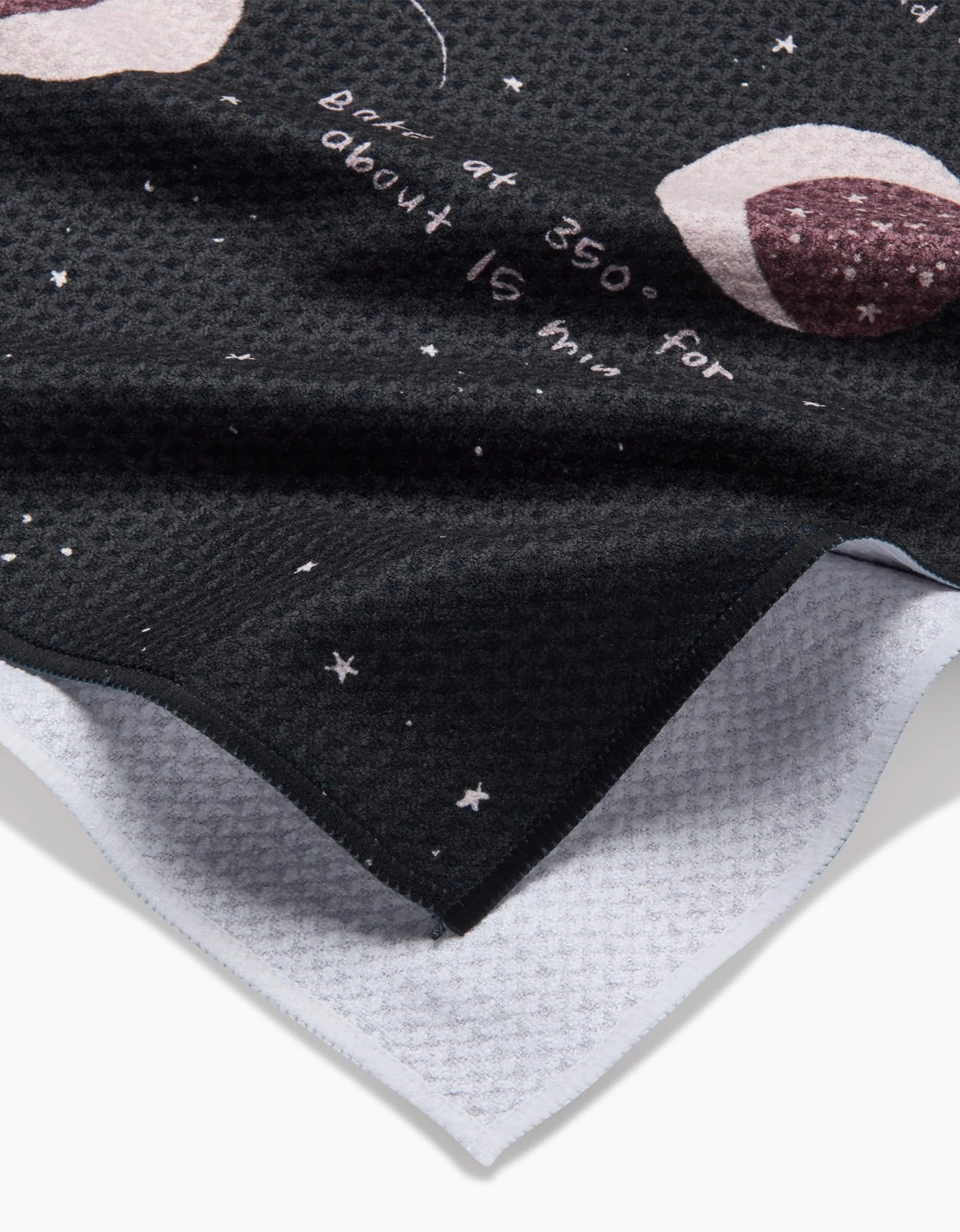 A close-up of the eco-friendly Full Moon Cookies tea towel by Janna Sue Design shows black textured fabric with white text, cookie illustrations, and stars. A folded corner reveals the absorbent white underside—ideal for any kitchen.