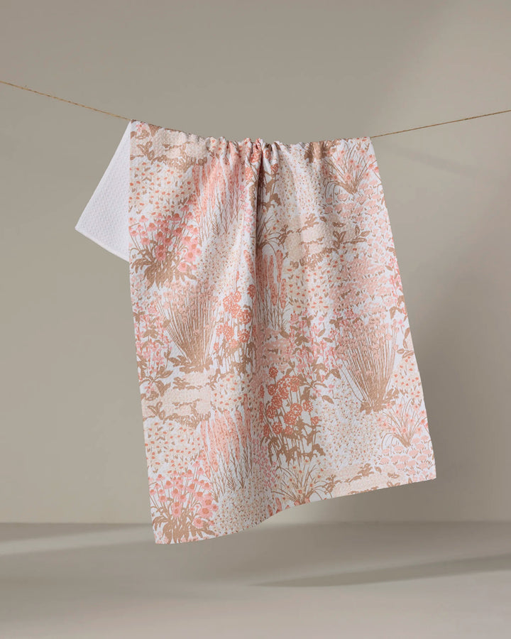 The Hufton Studio Garden Bloom tea towel, featuring a soft pink and beige floral pattern and made from recycled materials, hangs on a clothesline against a neutral backdrop.