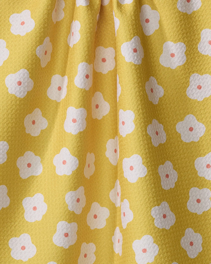 The Sara Mari "Garden Crush" kitchen towel features yellow textured fabric with a repeating white four-petaled flower pattern, each centered by a pink dot. Eco-friendly and stylish, it drapes softly, creating gentle folds.
