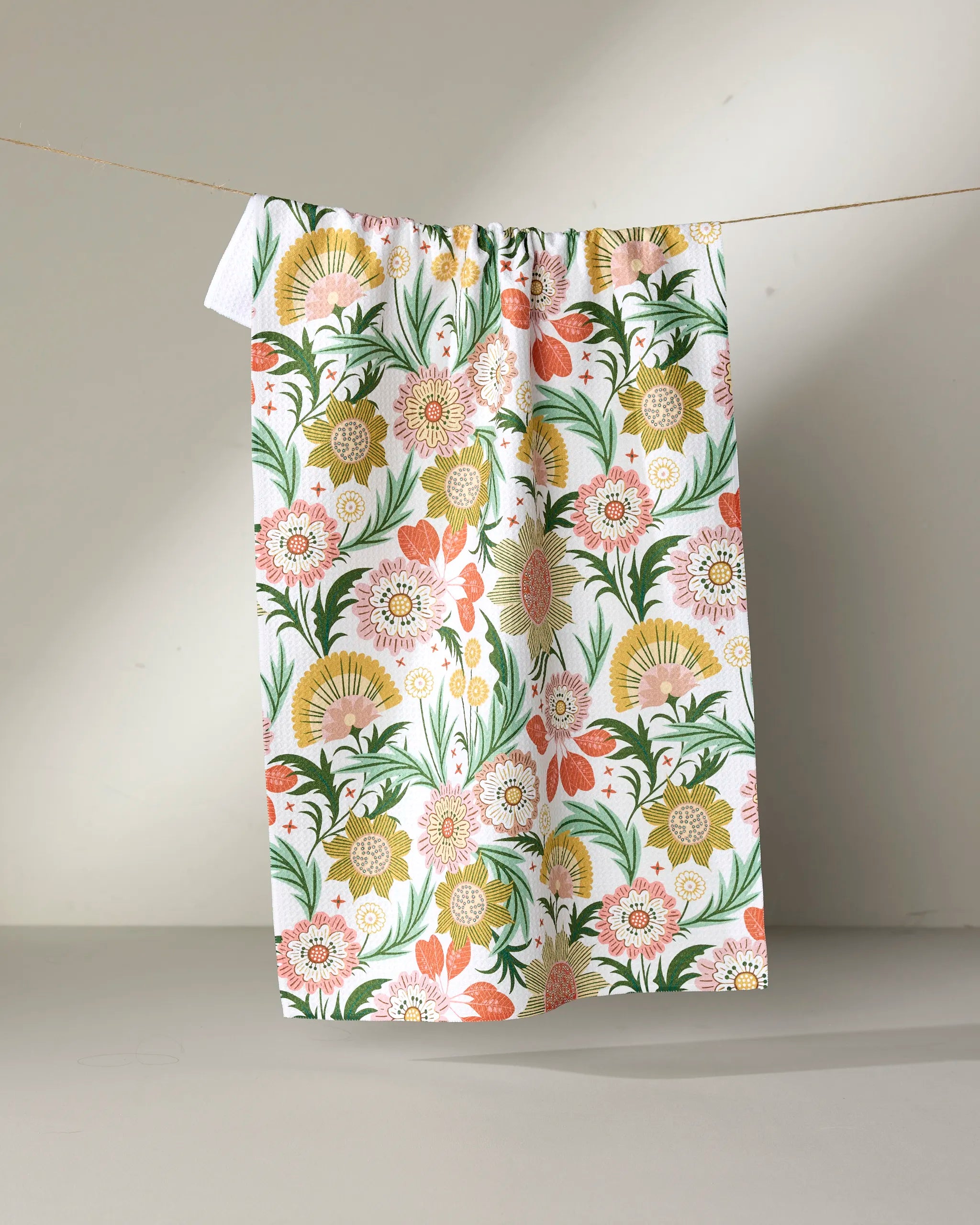 The "Garden Dreams" tea towel by Rachel Parker showcases a vibrant floral pattern with pink, yellow, orange, and green blooms and leaves, hanging against a neutral background.