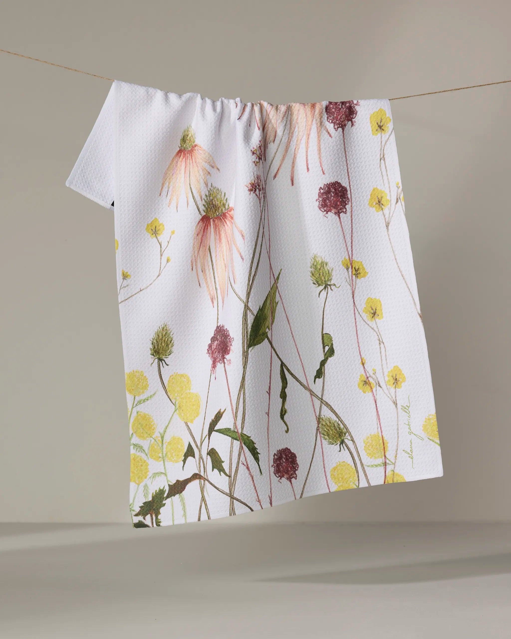 The Elana Gabrielle "Garden Flowers" kitchen towel, made from recycled materials, displays a vibrant floral pattern with yellow, pink, and green wildflowers as it hangs over a clothesline against a neutral background.
