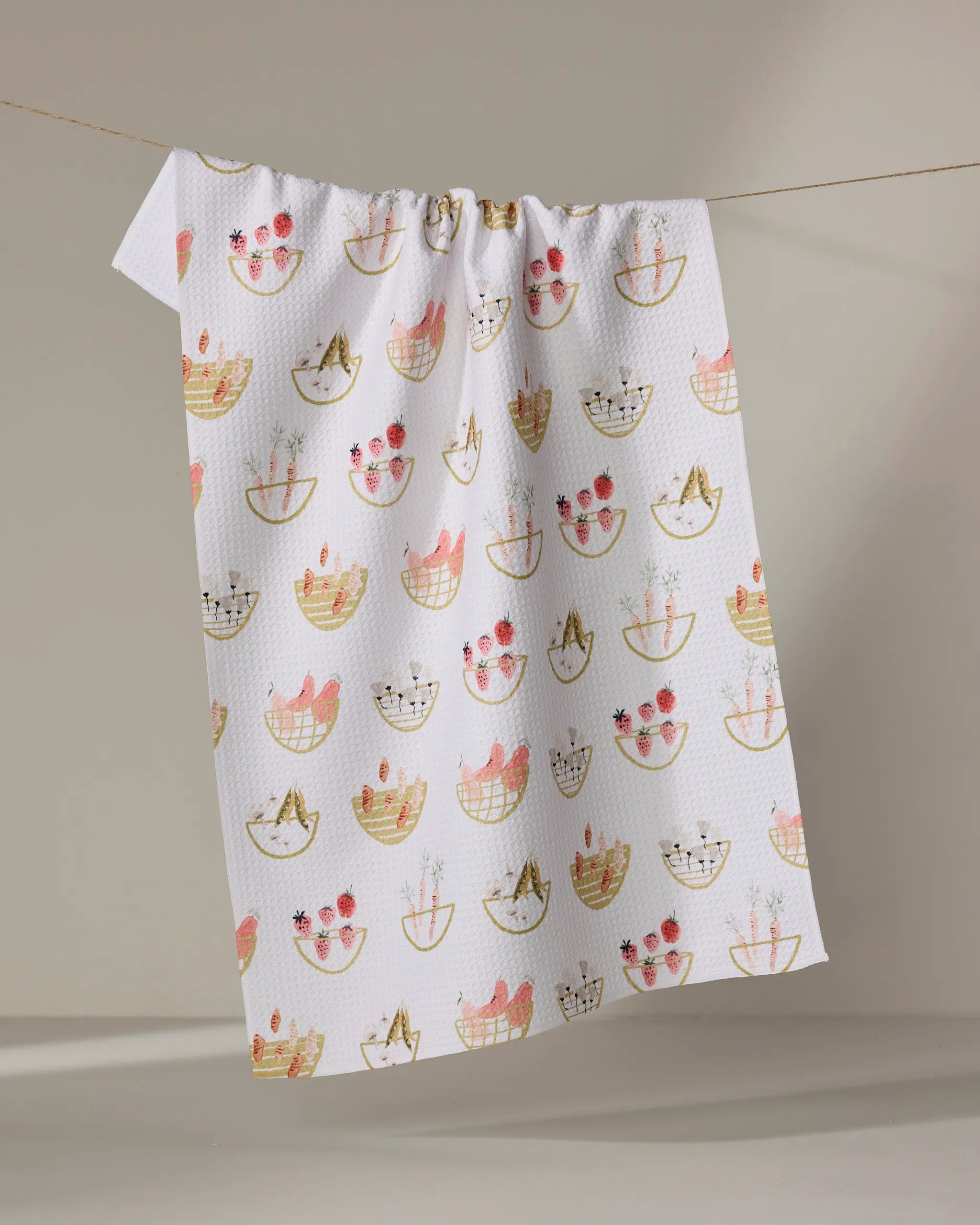 The "Garden Fresh" tea towel by Anee Shah is made from recycled materials and features a vibrant design of cats in baskets, potted plants, and flowers, shown hanging on a clothesline against a neutral background.