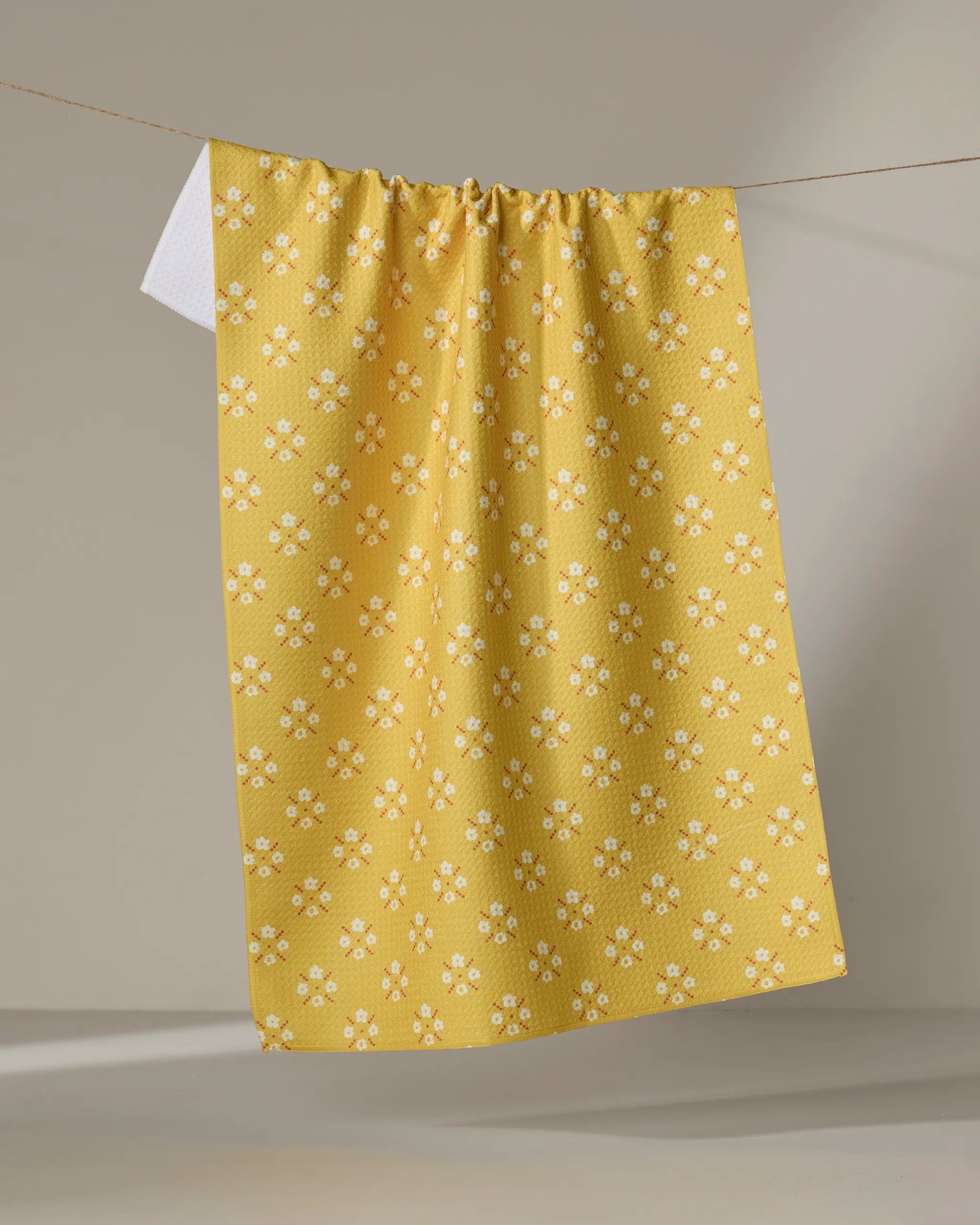 The Garden Lattice tea towel by Kelsey Weigl, featuring a yellow and white floral design and made from recycled materials, hangs on a clothesline against a neutral background.
