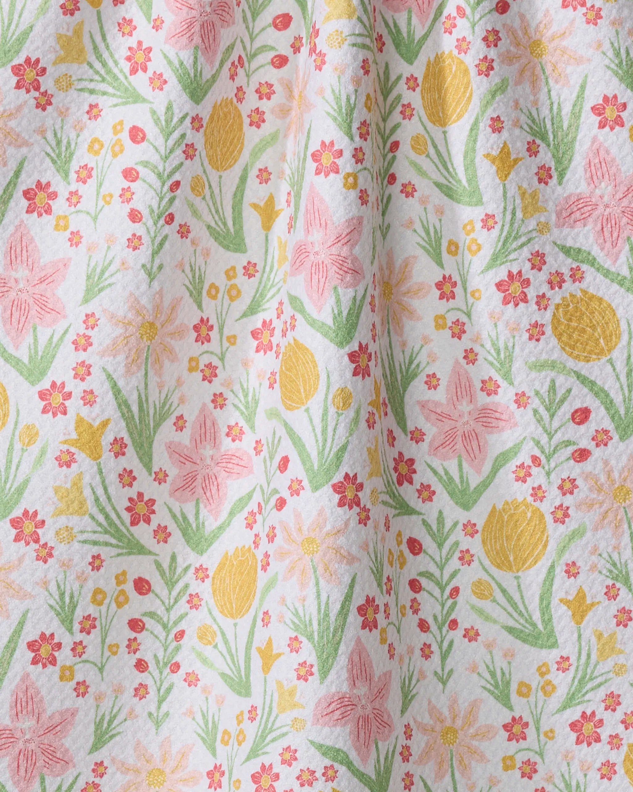 A close-up of the "Garden of Light" tea towel by Danelys Sidron, showcasing colorful yellow, pink, and orange flowers with green leaves on white fabric. Soft folds highlight its quick-drying quality.