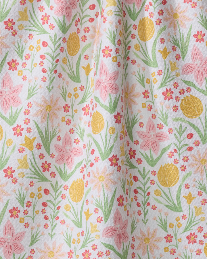 A close-up of the "Garden of Light" tea towel by Danelys Sidron, showcasing colorful yellow, pink, and orange flowers with green leaves on white fabric. Soft folds highlight its quick-drying quality.