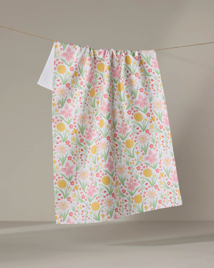 The “Garden of Light” tea towel by Danelys Sidron, featuring a quick-drying fabric and colorful pink, yellow, and green floral patterns, hangs on a clothesline against a neutral background.