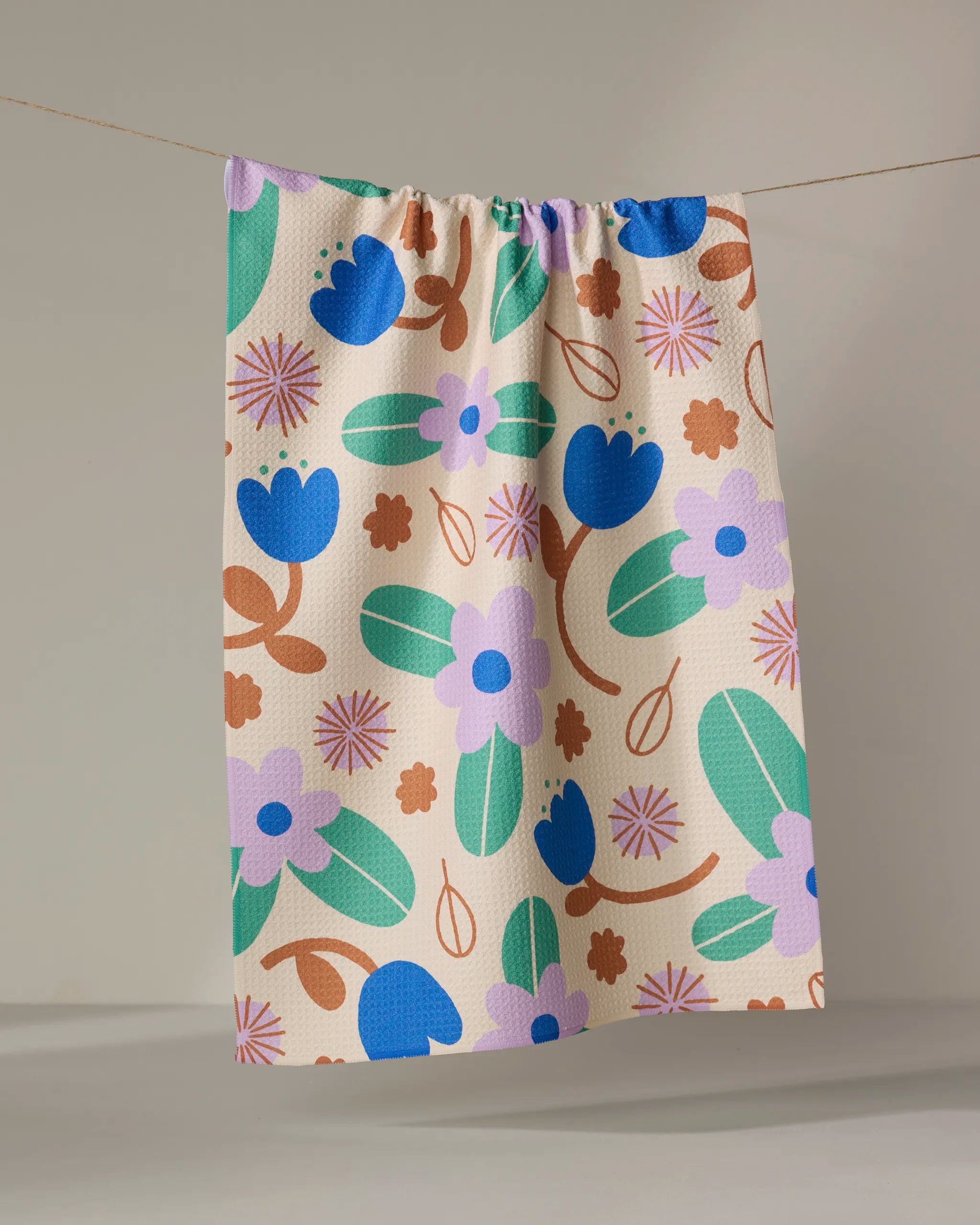 The Beetle Ink Co. Garden Patch recycled kitchen towel, decorated with blue, green, and brown floral patterns, hangs on a string against a plain light background.