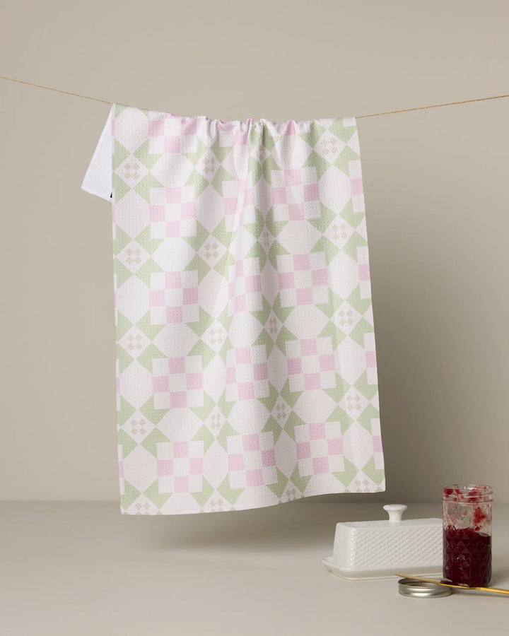 The Geometry Garden Quilt tea towel features pink and green geometric quilt designs, crafted from recycled materials, and hangs above a beige surface with a butter dish and a jar of red jam nearby.