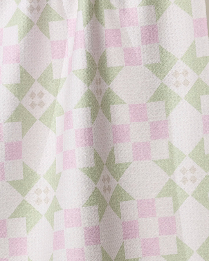 A close-up of the Geometry Garden Quilt tea towel shows its pastel pink and green checkerboard and diamond pattern on white, highlighting the absorbent, textured surface and gentle folds.