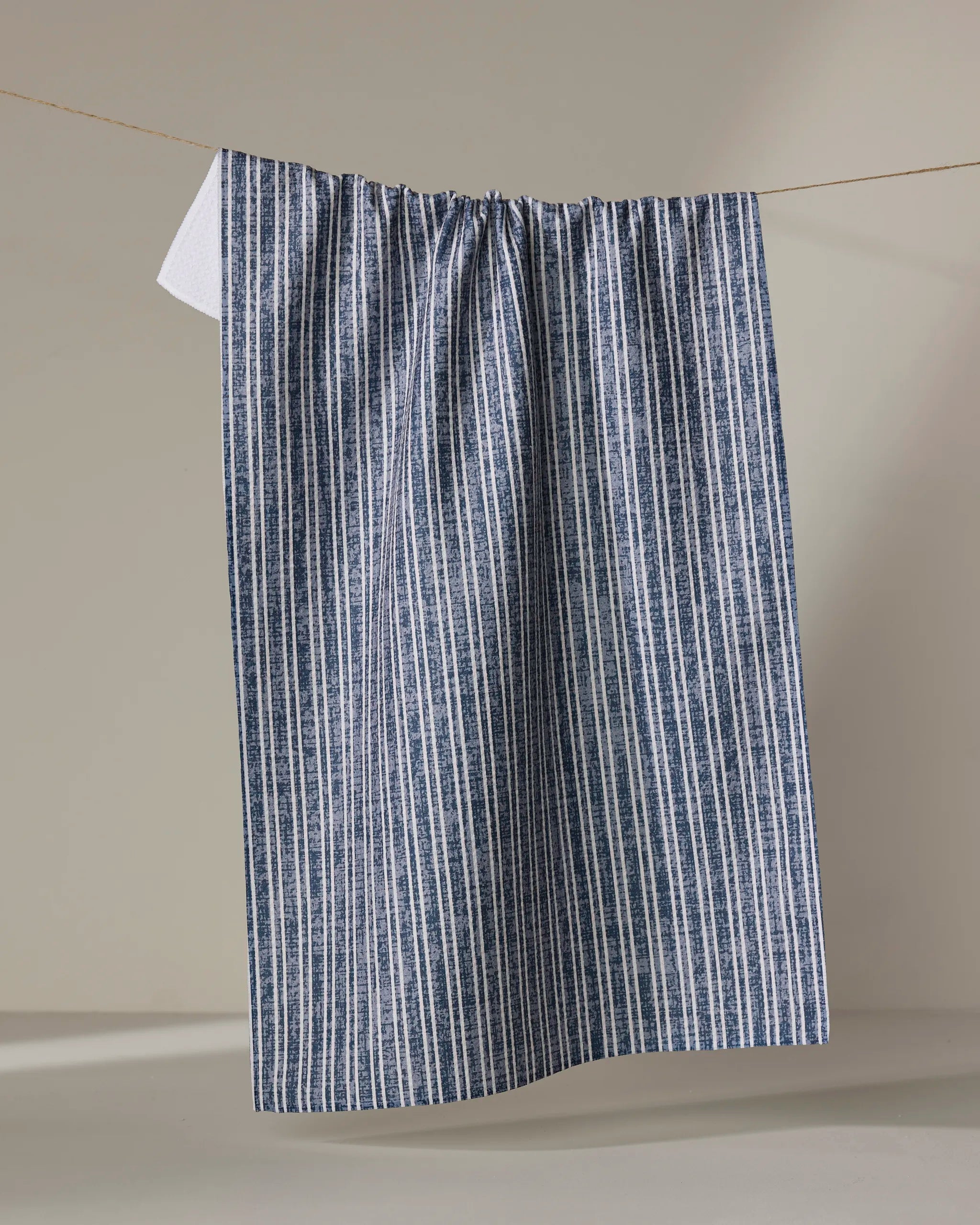 The Becki Owens Garff Blue tea towel, featuring blue and white vertical stripes and made from ultra-absorbent recycled materials, hangs on a clothesline against a plain beige background.