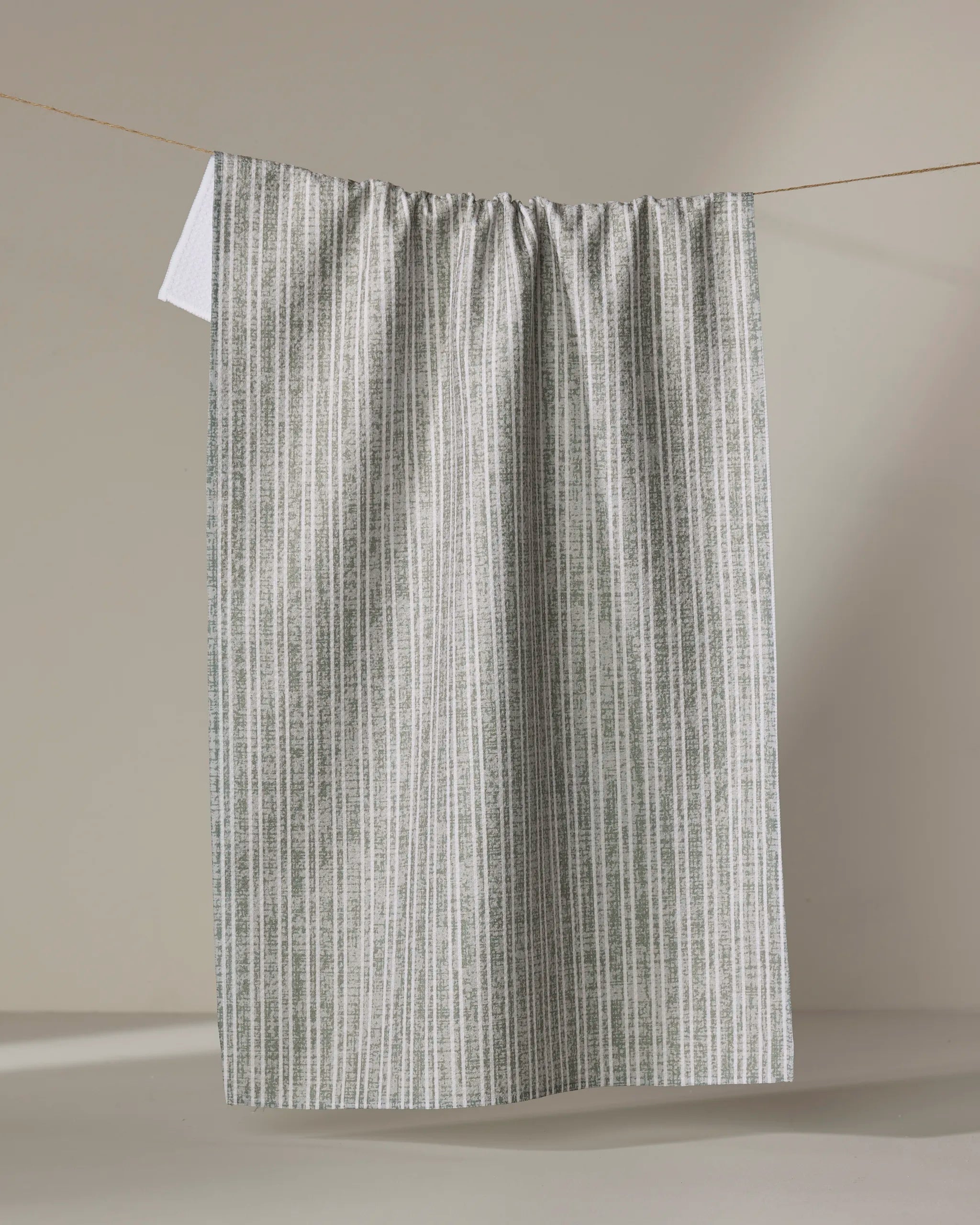 The Garff Sage tea towel by Becki Owens hangs from a clothesline against a neutral background. Its lightweight, eco-friendly fabric features subtle gray and white vertical stripes—perfect as an absorbent kitchen dish towel.