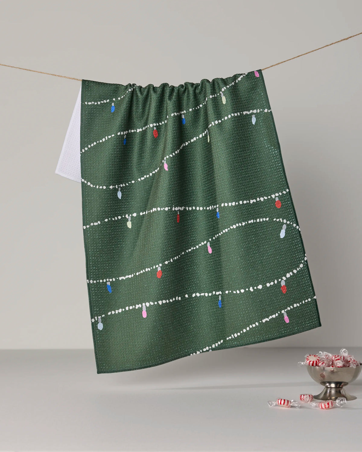 Geometry’s “Garland Glow” kitchen tea towel, crafted from recycled materials, features green fabric adorned with white dotted garlands and colorful light bulb designs. It hangs on a string with a white cloth partially visible behind it, while peppermint candies rest in a glass dish on the nearby white surface.