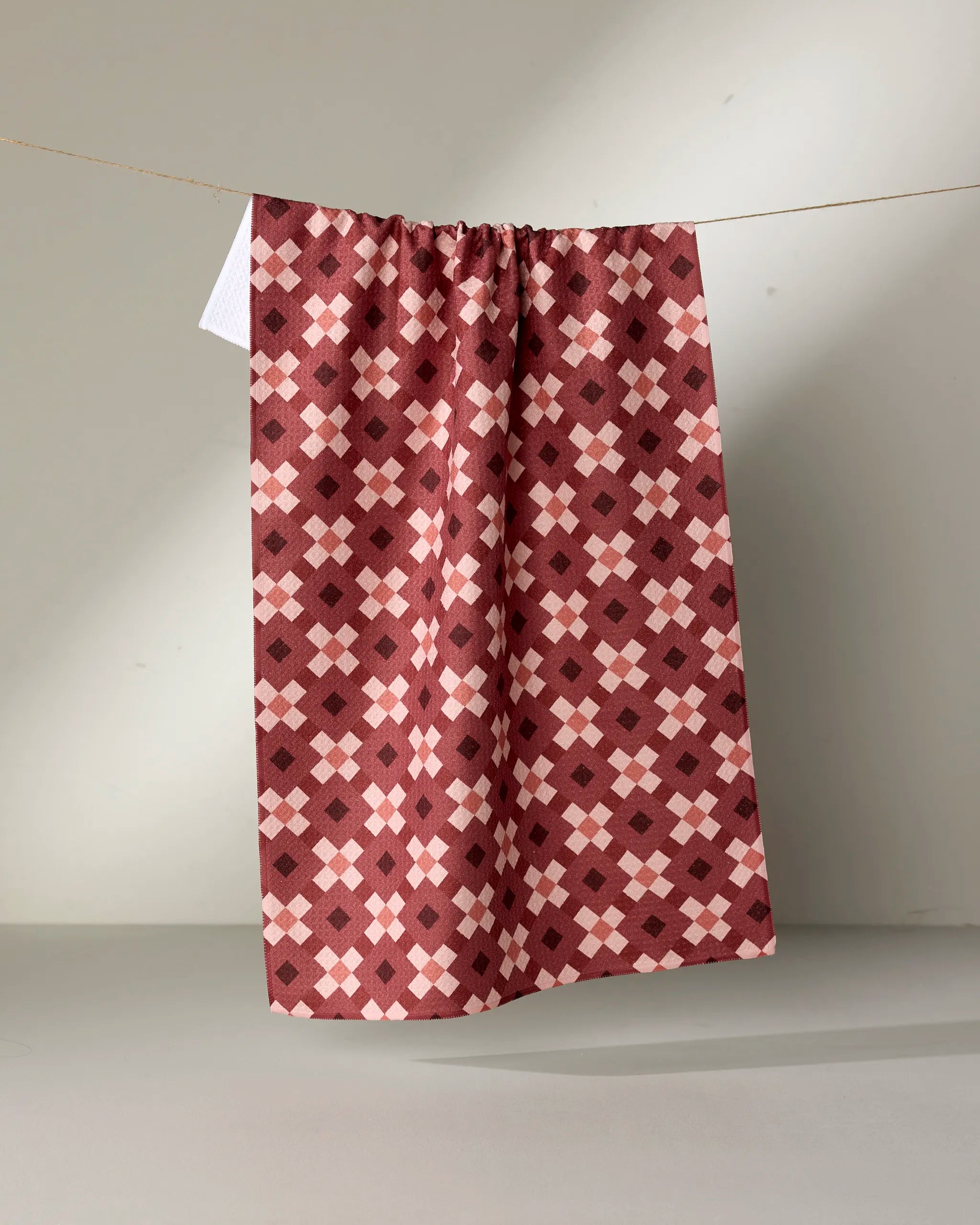 The Garnet Geo kitchen towel by Teri the Butter, made from recycled materials, displays maroon, pink, and cream geometric squares and diamonds and hangs on a line against a neutral background.