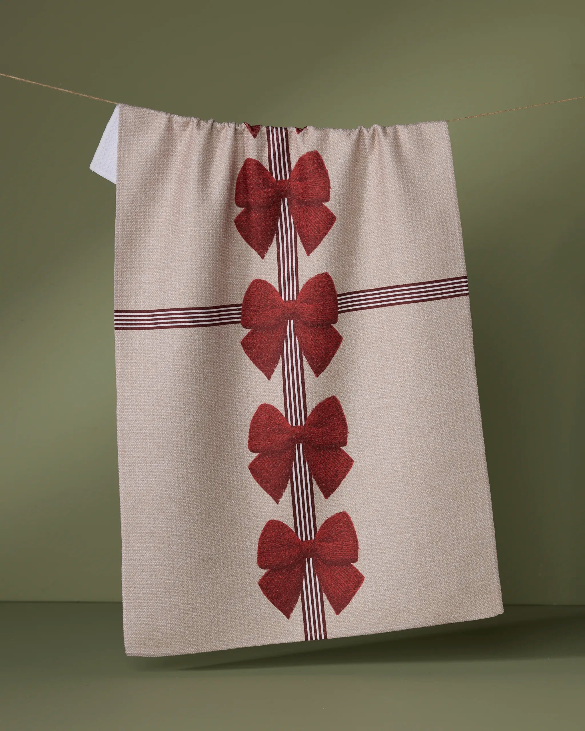 The Garnet tea towel by Geometry, made from recycled materials, hangs on a clothesline with red bows and thin red lines like a wrapped gift against an olive green background for a charming look.