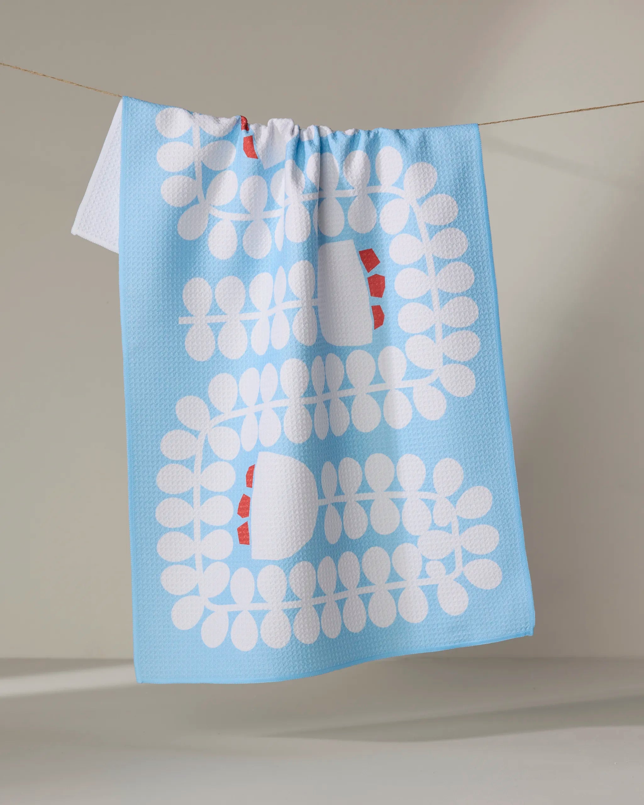 The Gemini kitchen towel by Happy Hemera, crafted from recycled materials, features a light blue color with a white geometric leaf pattern and red accents, hanging from a clothesline against a neutral background.