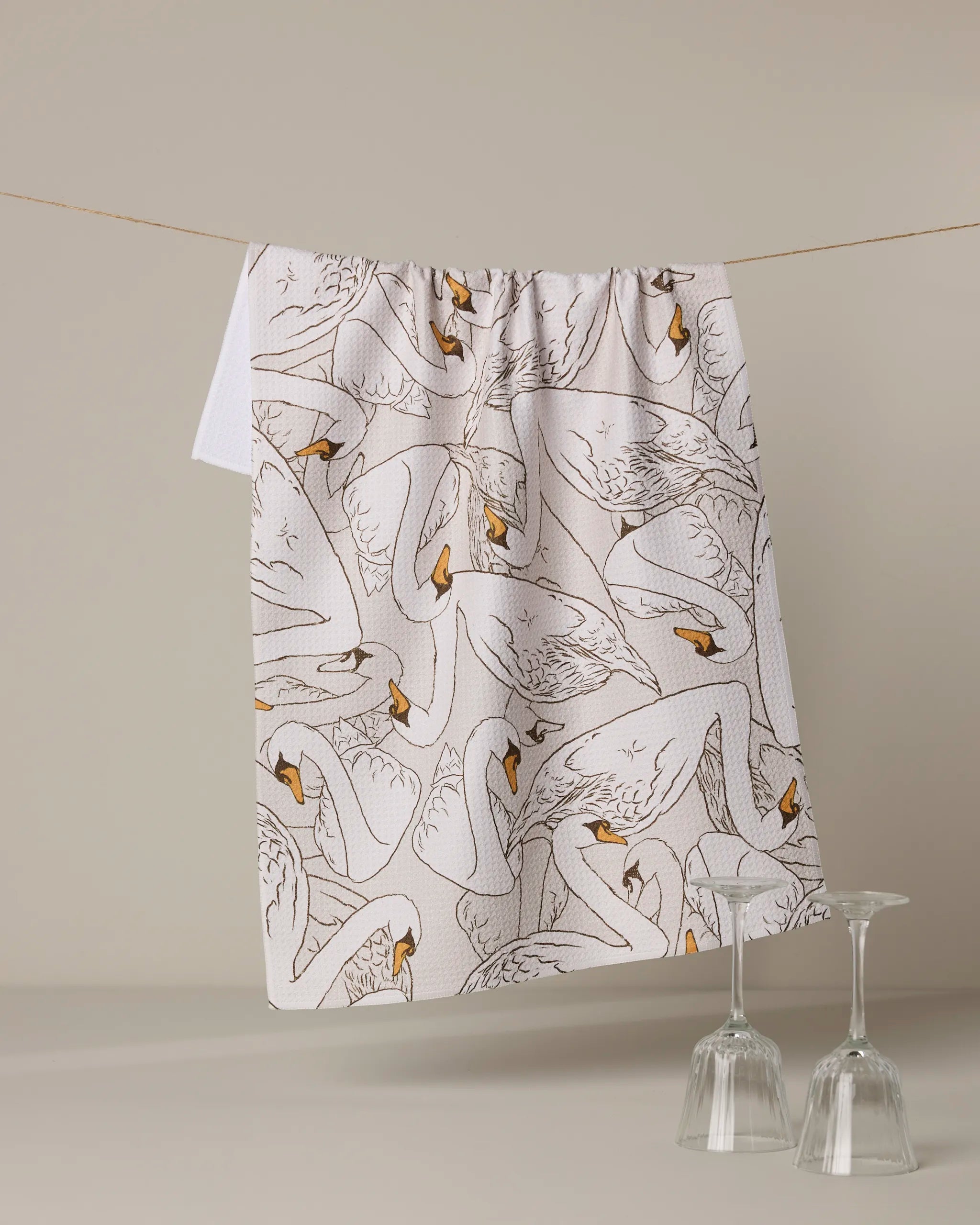 The Gentle Strength kitchen towel by Geometry, made from recycled materials and featuring an intricate swan design, hangs on a string above two upside-down wine glasses against a neutral background.