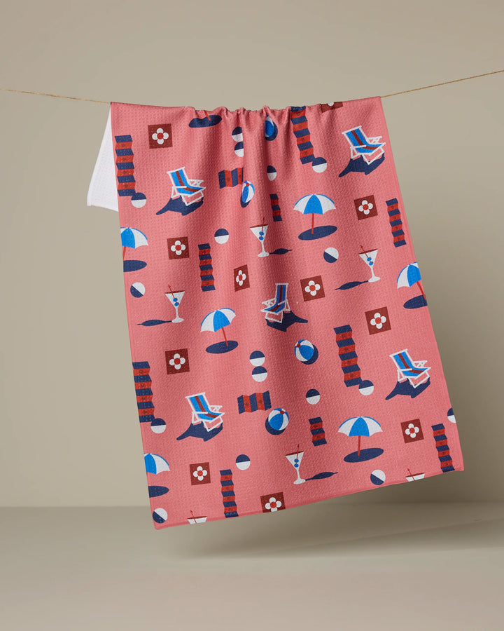 The Geometry Geo Beach Day tea towel, featuring playful summer icons in blue, white, and red, hangs on a line. Made from recycled materials, this cheerful pink towel brightens any kitchen.