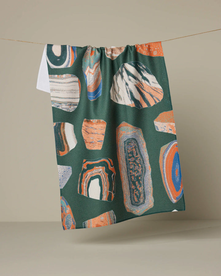 The Geo Class recycled kitchen towel by Geometry hangs from a line, displaying abstract orange, blue, white, and beige patterns that resemble sliced geodes or stones.